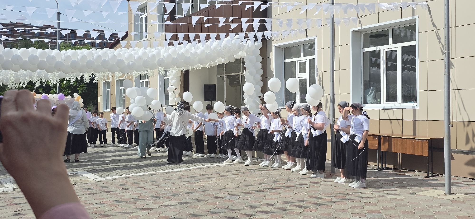 The image shows a group of children standing outside a building, likely a school, in an urban area. The location is marked by white flags and a string of white balloons that creates an archway over the area. The children are dressed in similar uniforms: white shirts and dark skirts or pants. Some of them hold white balloons. There are also adults present, guiding or organizing the children. In the foreground, there is an arm with a mobile phone camera extended, suggesting someone is capturing the scene. The background includes the building's facade with windows, a desk and visible architectural details.