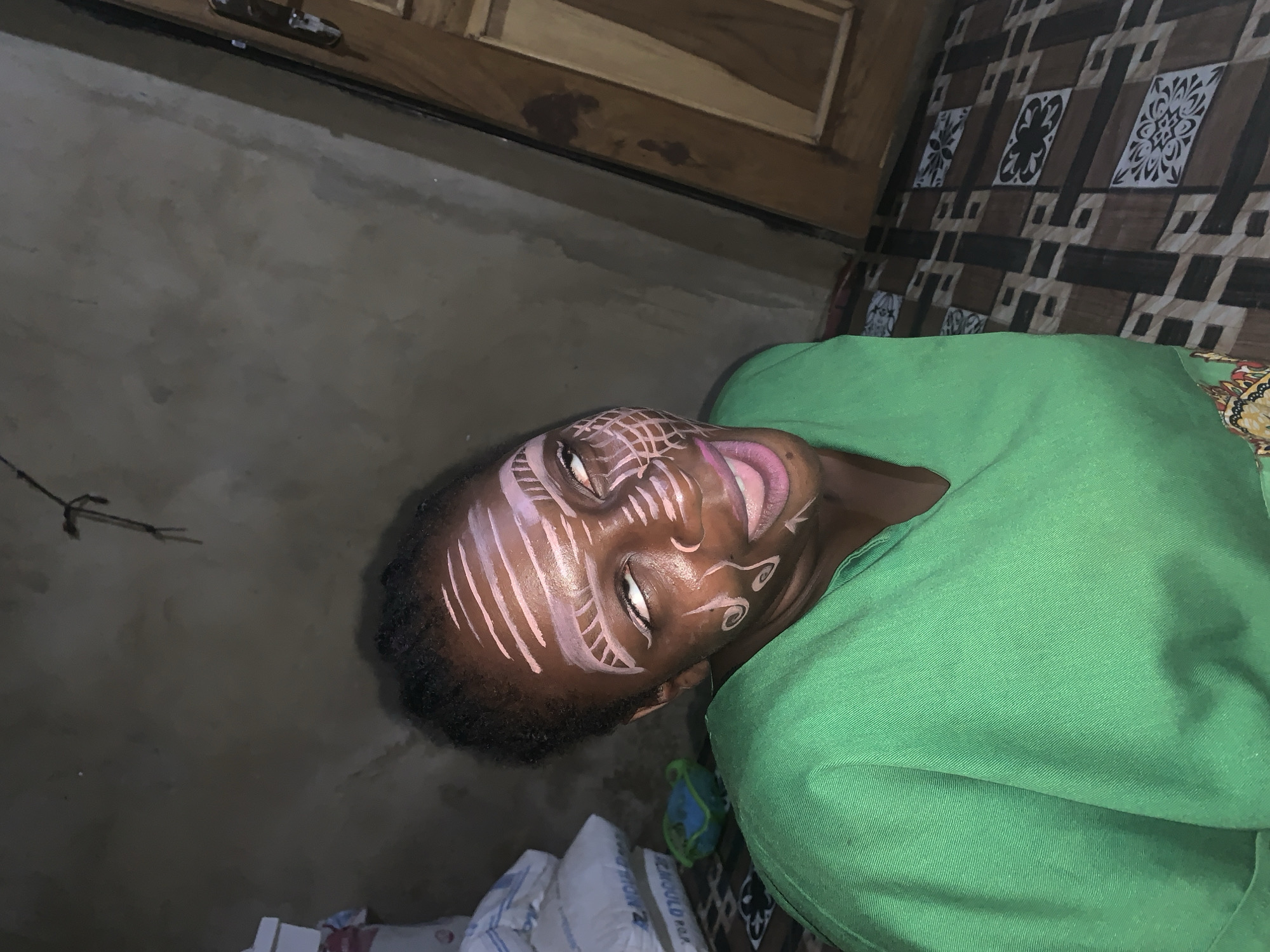 The image shows a young person with their face painted with intricate white and pink designs, lying down and wearing a green garment. The face paint includes stripes, curves, and circular patterns across the forehead, cheeks, and chin. The person's eyes are open, and their lips are slightly parted, with pink lipstick applied. The background consists of a textured wall, a wooden surface, and a patterned floor covering. There are also some white bags with visible text in the lower part of the image. The lighting appears to be artificial, suggesting an indoor setting. The overall mood seems to be relaxed or possibly contemplative, given the subject's pose and expression. The location is identified as Agban, Nigeria.
