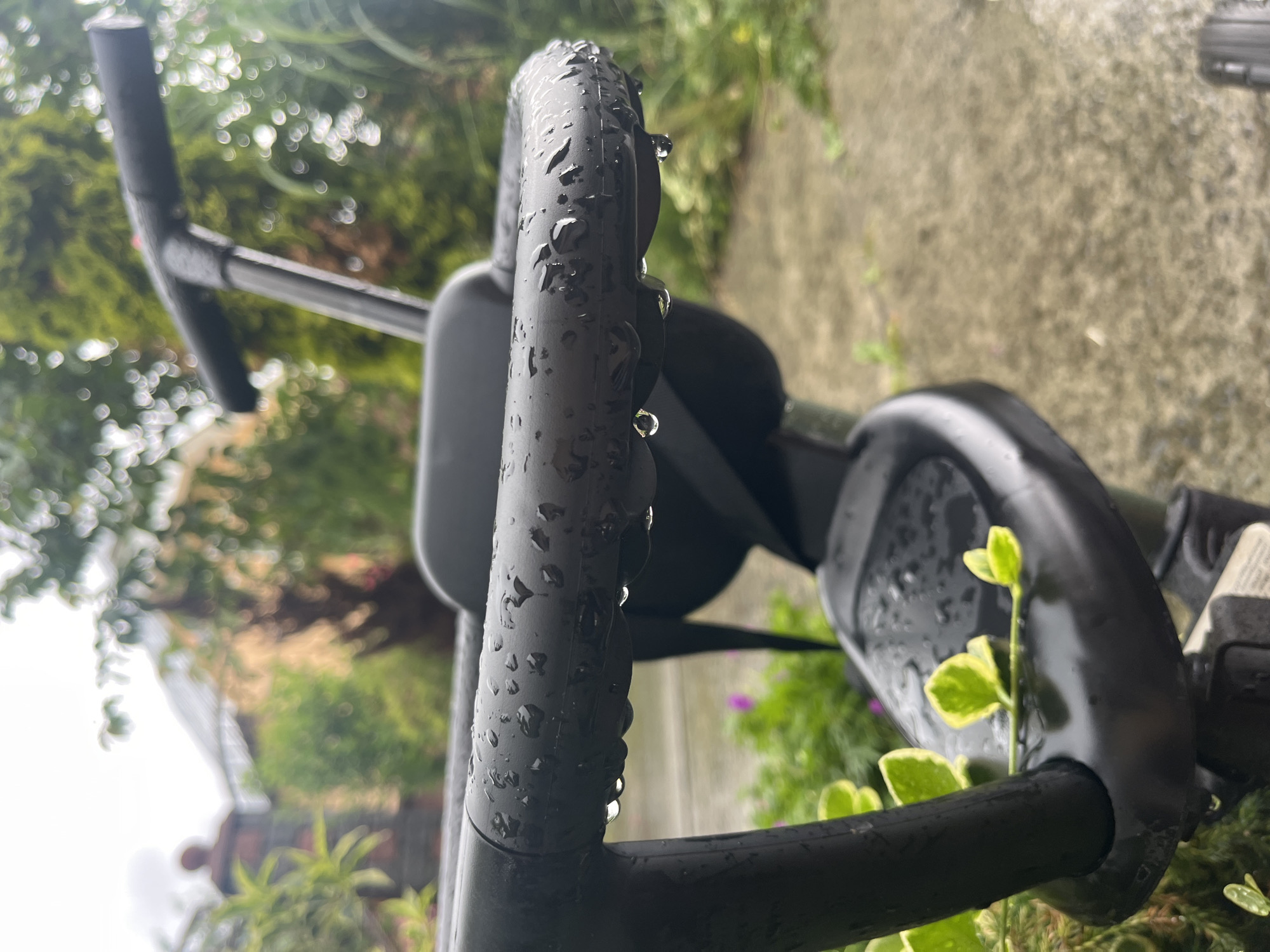 A close-up, low-angle shot focuses on the wet, black handlebar and seat of a child's tricycle. Water droplets cling to the curved handlebar, with several larger drops about to fall. The seat also shows water accumulation. In the background, out of focus, are lush green foliage, a paved surface that appears wet, and a hint of a building with a reddish-brown, figure-like shape on a balcony. The overall scene suggests an outdoor setting, possibly a yard or garden, likely after rainfall.