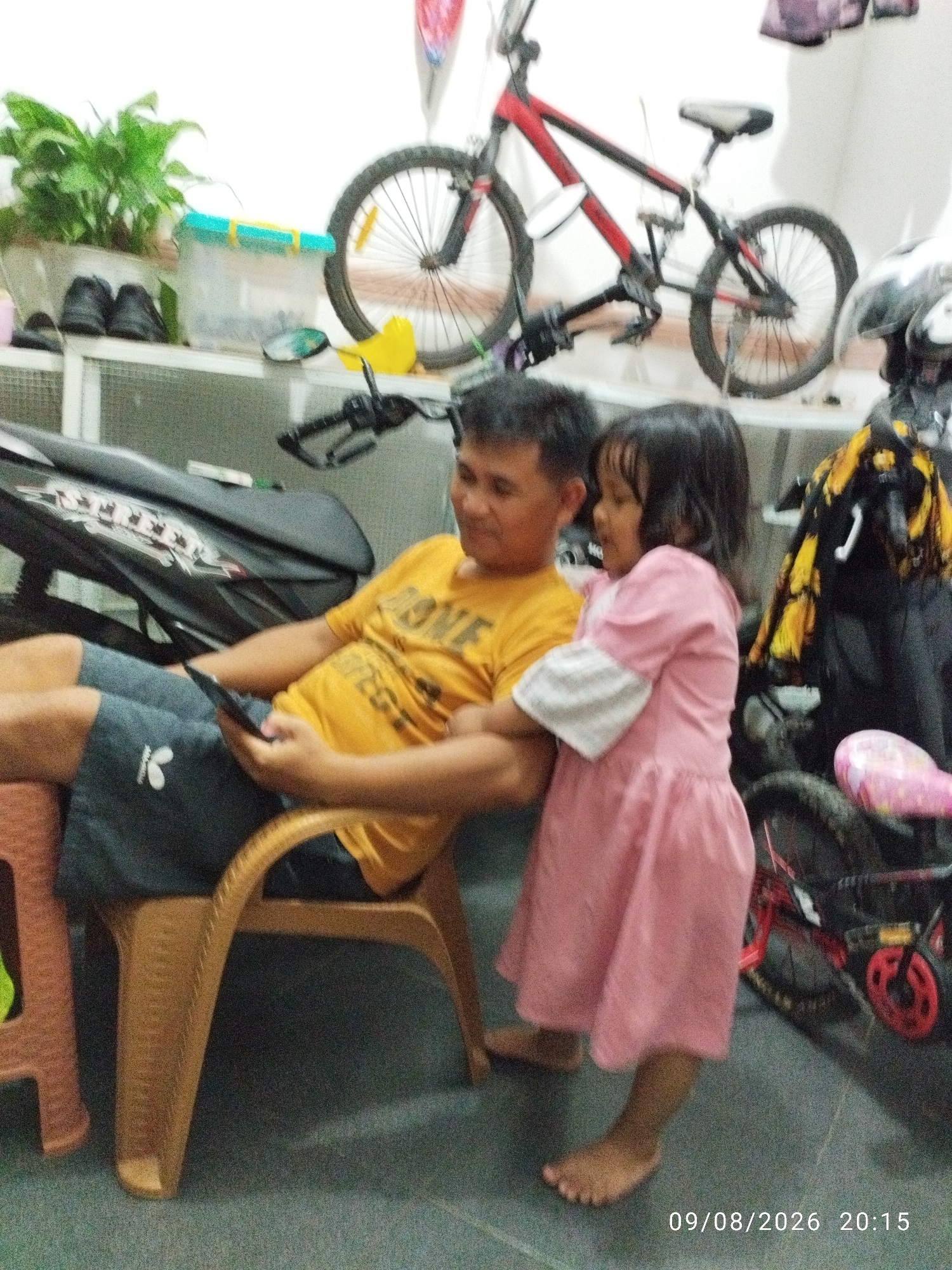The image depicts a father and his young daughter in what appears to be an indoor storage or garage area. The father, wearing a yellow t-shirt and dark shorts, is seated on a plastic chair and looking at a mobile phone. His daughter, dressed in a pink dress, stands beside him, looking up at him. In the background, various objects are visible, including bicycles, a motorcycle, a plant, and storage containers. The scene is illuminated by artificial light, suggesting it is either daytime with indoor lighting or evening. A timestamp in the bottom right corner indicates the date and time as 09/08/2026 20:15, placing the scene in the evening. The overall mood seems casual and domestic. There are no clear indications of weather conditions, but the setting suggests a residential or semi-commercial space in Bandar Lampung, Indonesia.