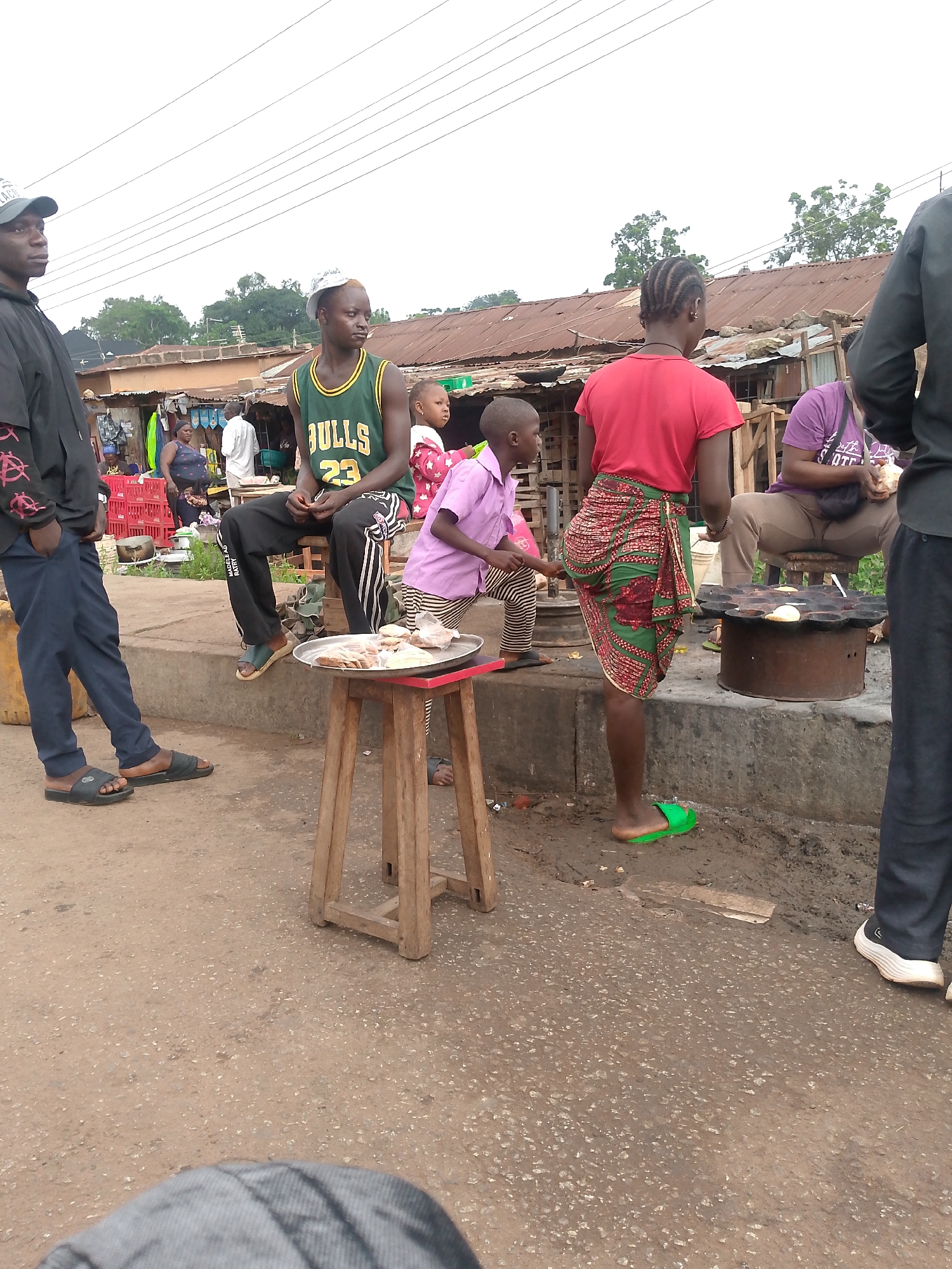 The image depicts an outdoor scene in Bukuru, Nigeria. Several people are present on what appears to be a street or roadside. A man in a black jacket and blue pants stands to the left. A young man wearing a "Bulls 23" jersey sits on a concrete ledge. A small child is standing by a wooden stool with a tray of what might be food items on it. A woman in a red shirt and patterned skirt stands near a circular metal cooking device, where another woman in a purple shirt is seated. A man in black is standing to the right, partially cut off in the frame. In the background, there are buildings with corrugated iron roofs, red plastic crates, and other people. Overhead, power lines are visible. The environment appears to be somewhat urban and possibly a commercial area with street vendors.