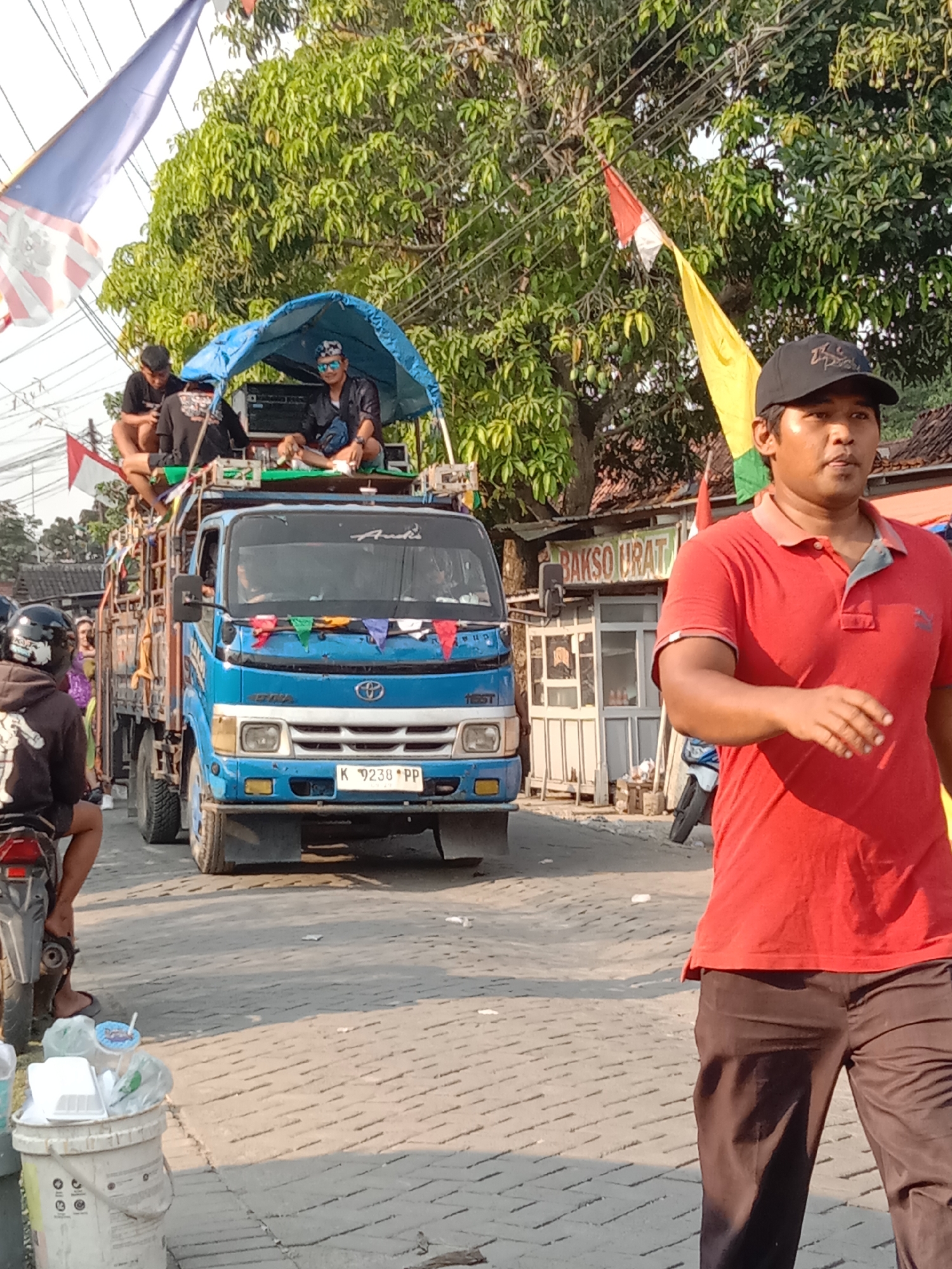In Wirosari, Indonesia, a blue truck with people and equipment is driving down a paved street. The truck has a canopy on the back, covered with a blue tarp, and several people are sitting or standing on it. One person is wearing a black baseball cap and a red polo shirt, walking in the foreground. In the background, there is a building with a sign that reads "BAKSO URAT". Trees and flags are visible, suggesting a festive or celebratory event. Several white plastic containers filled with trash are on the side of the road. The street is lined with trees and electrical wires. The overall scene suggests a procession or parade.