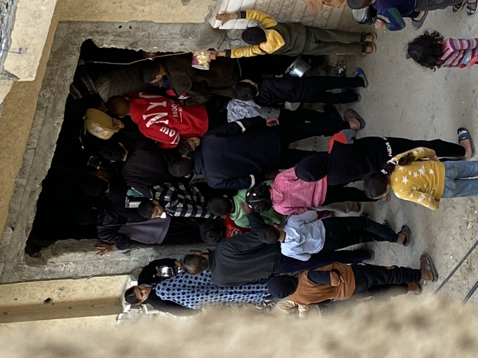 A crowd of approximately 25 individuals, predominantly children and several adults, congregates in an unpaved outdoor area adjacent to a dark, irregularly shaped doorway within a light-colored building in Khan Yunis, Palestinian Territory. The doorway features rough concrete framing, suggesting a breached or unrefined entry point. Individuals are tightly packed, facing towards and partially entering the dark interior.
On the left, a woman in a black top and a blue and white patterned long dress stands, holding a small metallic container. An adult male with dark hair and a dark sweater is visible foreground-center, facing away. The majority are children of varying ages, dressed in casual attire including hoodies, sweaters, and long pants. Notable garments include a red hoodie with "NYC BROOKLYN" text, a yellow hoodie, and a black and white striped shirt.
Several individuals, particularly children, are holding up metallic bowls, pots, or packaged items. A child in a yellow hoodie on the right has arms raised, holding a metallic pot and a small yellow-orange packaged item against the wall. A large, circular, light-colored basin or tray is visible within the dark opening, held by an unseen person. The scene depicts a congested gathering at what appears to be a point of resource distribution or access.