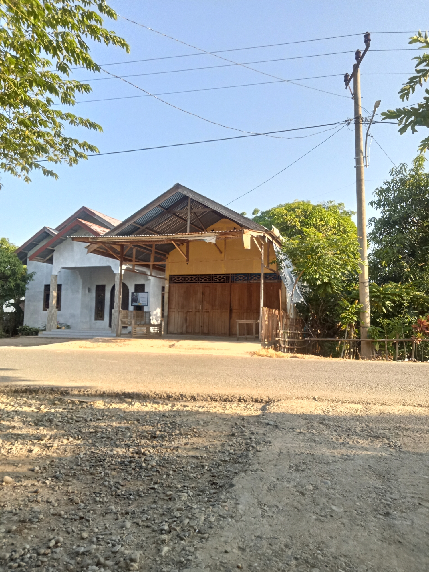 The image depicts a street view in Matangsijuek Timur, Indonesia. In the foreground, a paved road runs horizontally across the frame, with a gravel shoulder on either side. Beyond the road, a two-story building is visible, with a light-colored facade on the left and a yellow section on the right. The building has a corrugated metal roof supported by wooden beams. The yellow section features large wooden doors, suggesting a shop or garage. A small, framed sign is placed on a wooden bench in front of the building. To the right of the building, a concrete utility pole with electrical wires is present, along with lush green trees and vegetation. The sky is clear and blue, indicating daylight.