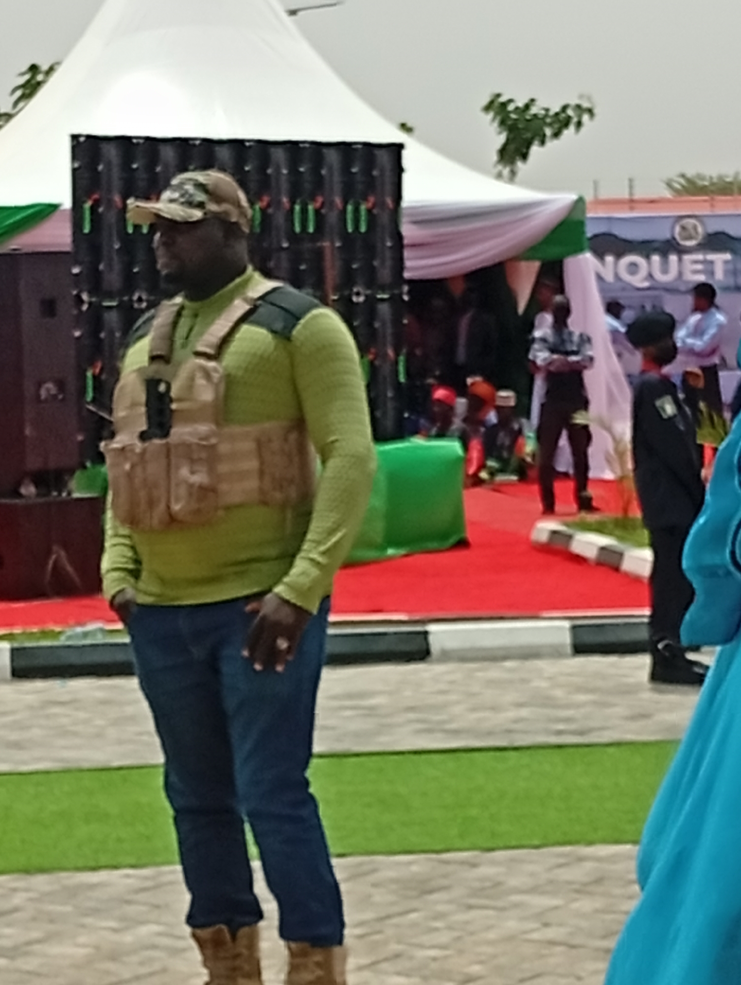 The image depicts an outdoor event, likely a banquet or public gathering, set in Unguwan Sanusi, Nigeria, during daylight hours under an overcast sky.
In the foreground, a man stands watchfully, facing left. He wears a light green long-sleeved shirt, dark blue jeans, and brown boots. Over his shirt, he has donned a tan tactical vest, and his head is covered with a camouflage baseball cap. A prominent ring is visible on his left hand, which is casually placed in his pocket.
The background reveals the event's setup: two large white tents, one with white and green draping, the other with white and pink, are erected over a red carpet. To the left, a stage or backdrop comprises black panels decorated with green and red accents, with large speakers positioned on either side. Numerous individuals are scattered throughout the background; some are dressed in light-colored shirts, while at least one person is seen in a dark uniform with a beret.
A low, striped barrier (black and white) delineates a strip of green grass from a paved section in the midground. On a blue banner or sign in the distance, the partial text "NQUET" is visible, indicative of the word "BANQUET," alongside a circular logo. The scene suggests a secured or organized public event.