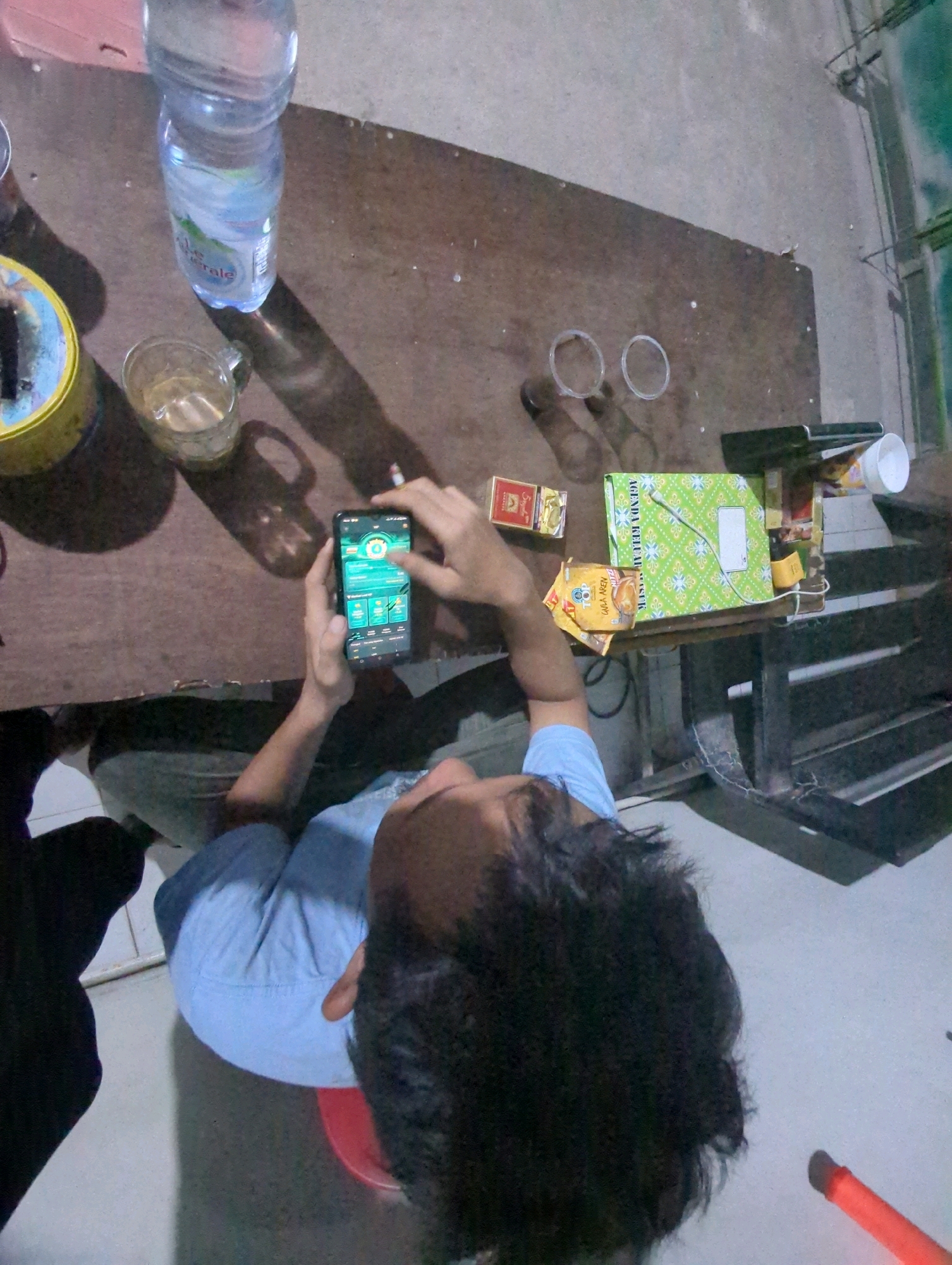 The overhead, high-angle shot depicts a dimly lit room, likely a simple dwelling in Batu Belaman, Indonesia, with objects spread across a brown wooden table. In the foreground, a person is seated, looking down at a smartphone displaying a green interface. Their left hand is holding the phone while their right hand is near the screen. The person wears a light blue short-sleeved shirt and dark pants.
To the left of the person, a large plastic bottle of mineral water is positioned. Next to it, a clear glass contains a light-colored liquid. To the right of the water bottle, a small yellow container with a blue rim is visible.
Further to the right on the table, a green box with a floral pattern is placed, alongside two small red boxes that appear to be cigarette packs. Two small, clear plastic cups are also on the table, along with a yellow object resembling a small charger.
The floor appears to be a light gray concrete. Shadows are cast across the table and floor, indicating a light source from above and slightly to the left. The overall impression is one of casual activity, possibly a gathering or downtime, in a straightforward living space.