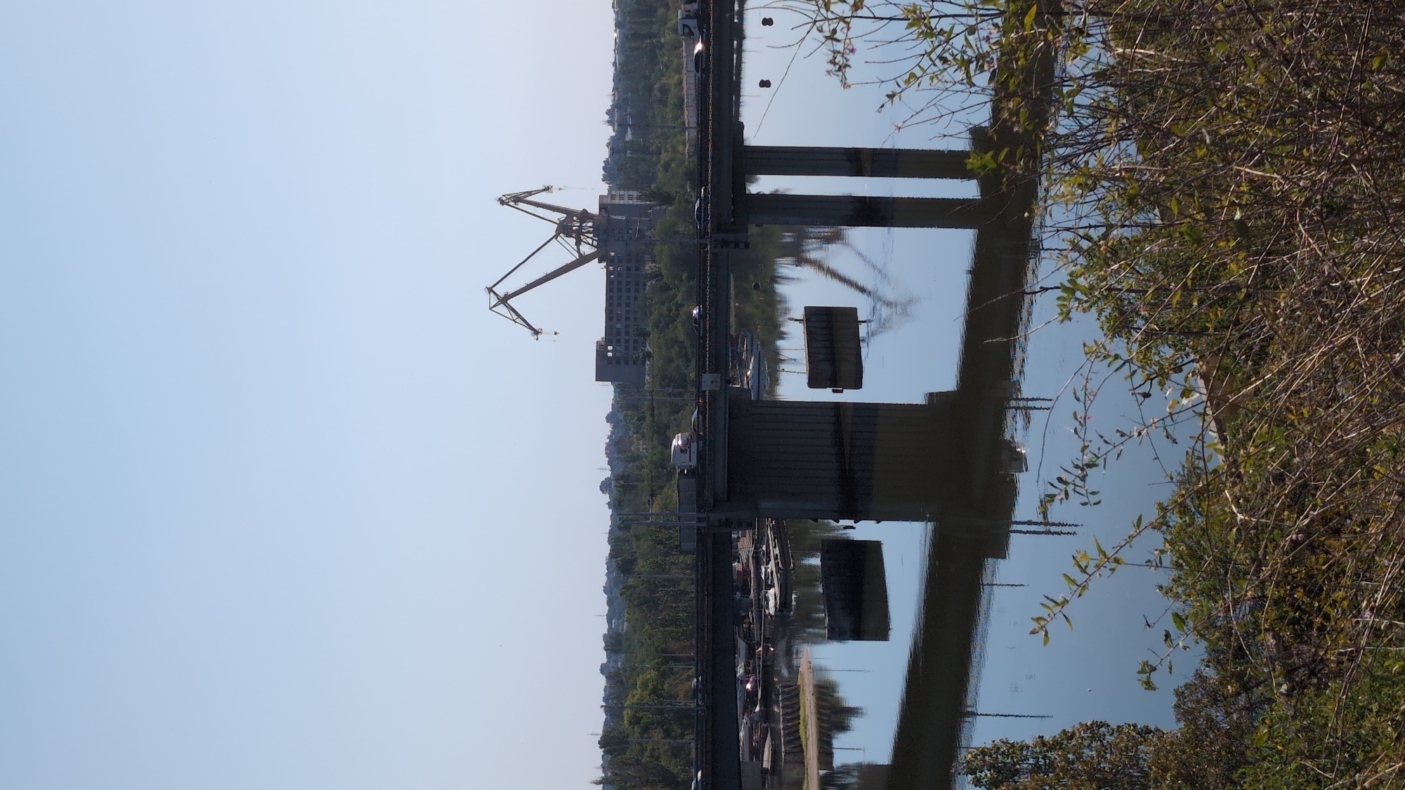 The image captures a wide shot of a river in Mykolaiv, Ukraine, with a multi-level bridge spanning across it. Two massive industrial cranes are visible in the background, with one of them partially obscuring an unfinished, multi-story building. Several vehicles, including trucks and cars, are actively crossing the bridge, indicating ongoing traffic. Various boats and barges are docked or moored along the riverbanks. The water's surface reflects the bridge structures and the cranes, creating a symmetrical visual effect. Dense green foliage lines the far bank of the river, and in the foreground, dry branches and bushes are prominent. The sky is clear and blue, suggesting a sunny day.