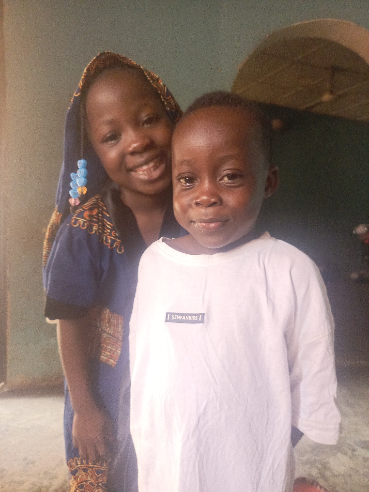 Two dark-skinned children are pictured indoors. The child in the foreground wears a white t-shirt with the label "SENFANKIDS" on the left chest. The child behind is wearing a blue patterned garment and a head covering adorned with beads. The location is likely Akwanga, Nigeria. An indoor setting with neutral colored walls can be seen. A ceiling fan is visible in the background. The children are smiling slightly.