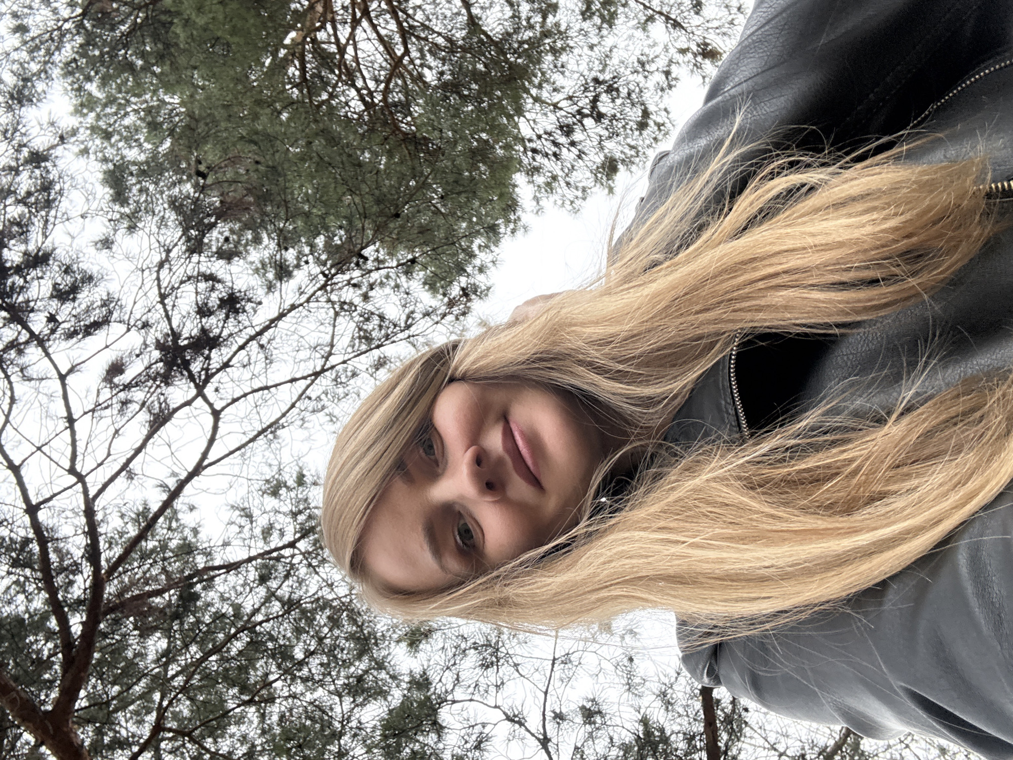 The rotated image features a young woman captured from a low, upward-looking perspective, set against a backdrop of trees and an overcast sky. She has long, straight blonde hair that cascades around her face and over her shoulders. She is wearing a black leather jacket, visible on her torso and arm. Her face is angled towards the camera, displaying light-colored eyes and a neutral to subtly pleasant expression with slightly parted lips.
The background consists of a dense canopy of trees against a bright, diffused sky, suggesting an overcast daytime. There is a mix of bare, intricate branches of deciduous trees and the dark green needles of evergreen pine trees, indicating a natural outdoor environment like a forest or park. The presence of both bare and evergreen trees suggests a colder season such as late autumn, winter, or early spring in Chaikovsky, Russia.
The image appears to be a selfie or a close-up portrait taken with the camera pointed upwards towards the woman, creating a dynamic and unusual viewpoint. No visible text is present in the image.