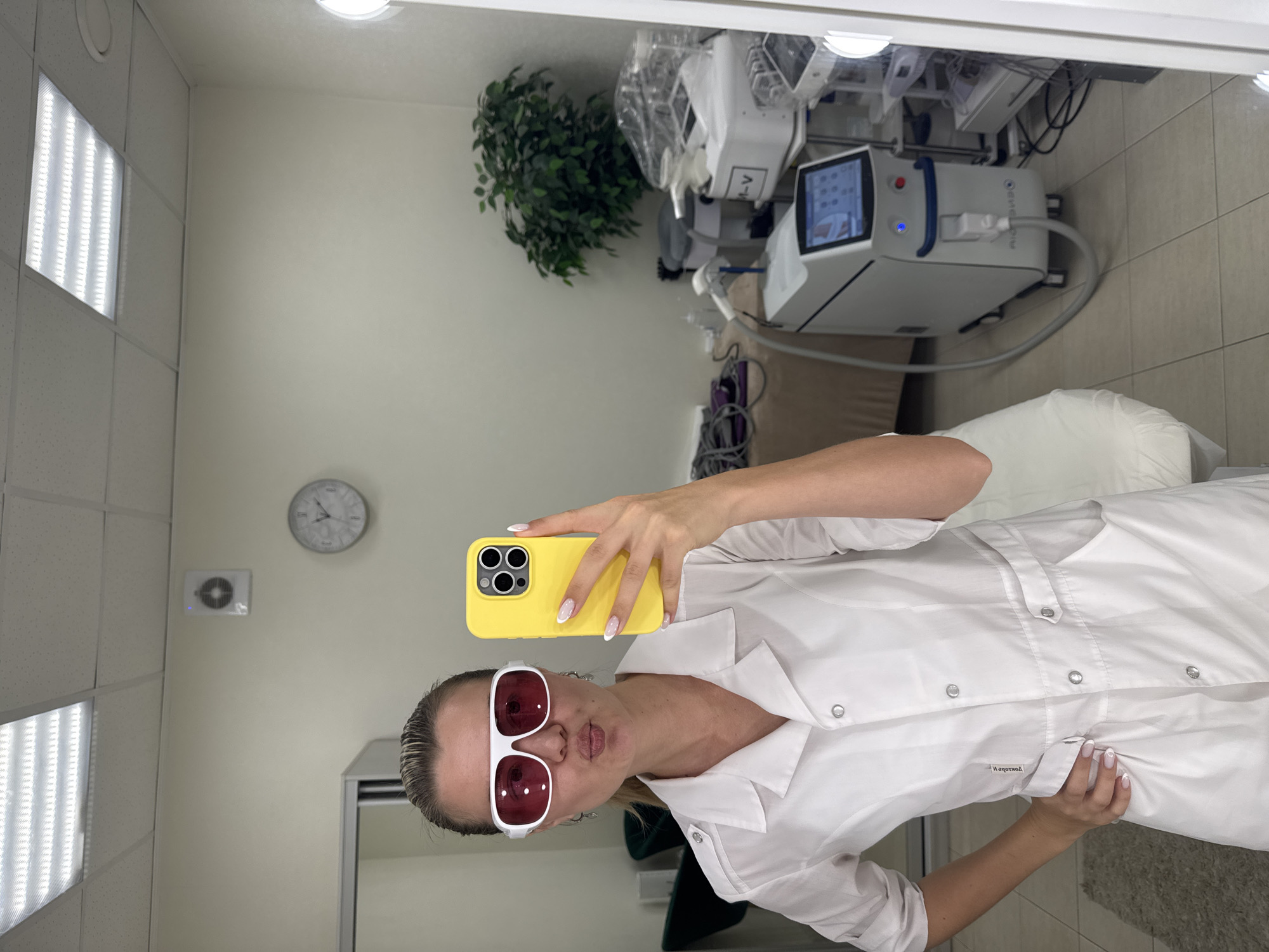 A mirror selfie in Unknown, Unknown shows a person in a white coat holding a yellow phone. The person wears white sunglasses with red lenses. Behind the person is medical equipment, a plant, a clock on the wall, and overhead lighting.