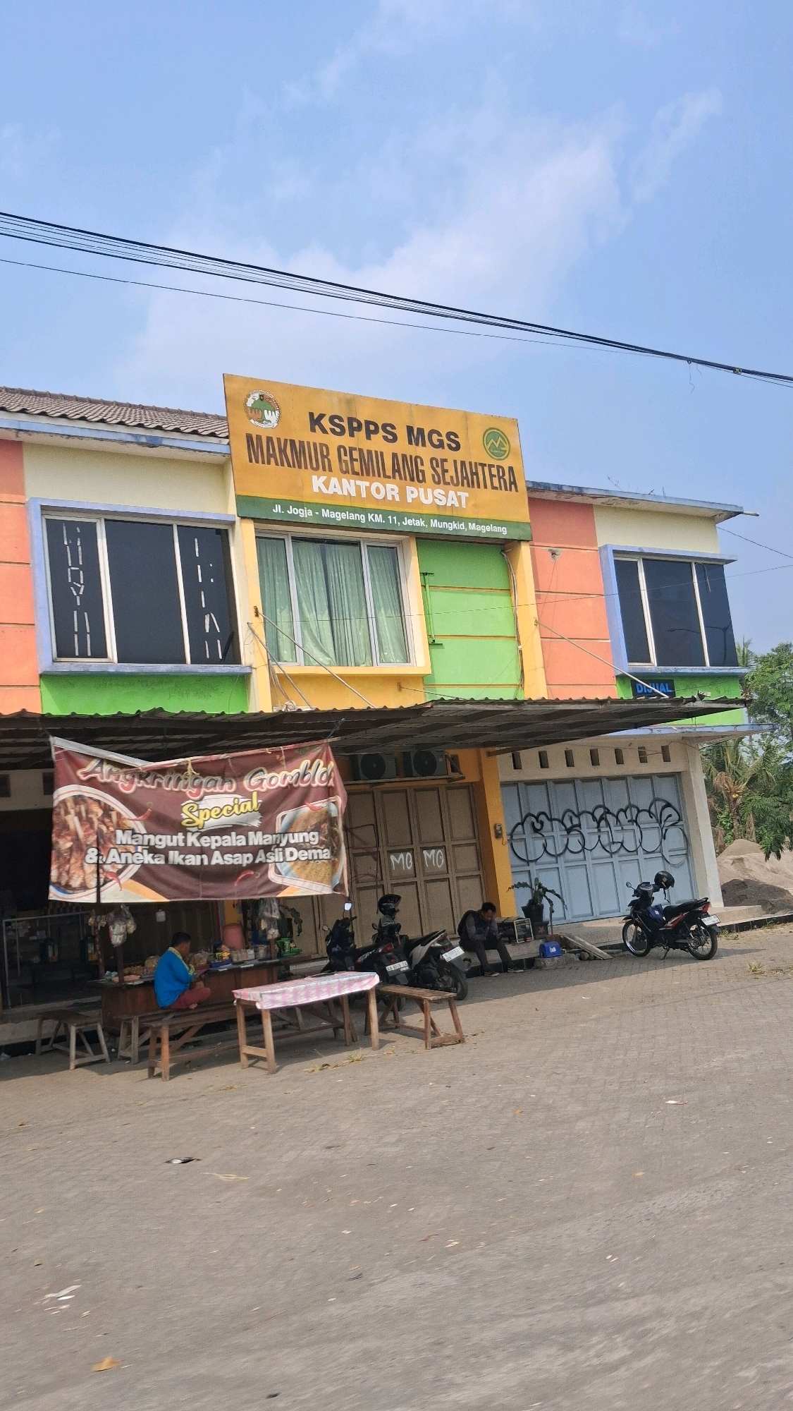 The image shows a building with multiple colored sections and a prominent yellow sign at the top. The sign reads "KSPPS MGS MAKMUR GEMILANG SEJAHTERA KANTOR PUSAT" and below that, "Jl. Jogja - Magelang KM. 11, Jetak, Mungkid, Magelang". This indicates the location is on the Jogja-Magelang road, kilometer 11, in Jetak, Mungkid, Magelang. The building has large windows and a covered area in front. A banner hangs from the covered area, advertising "Angkringan Gomblong Special" with images of food. Beneath the banner, a small food stall is visible with a person sitting at it. Several motorcycles are parked in front of the building, and two people are sitting on a bench in front of the motorcycles. The environment appears to be an urban or semi-urban setting, with a road in the foreground and a clear sky above.