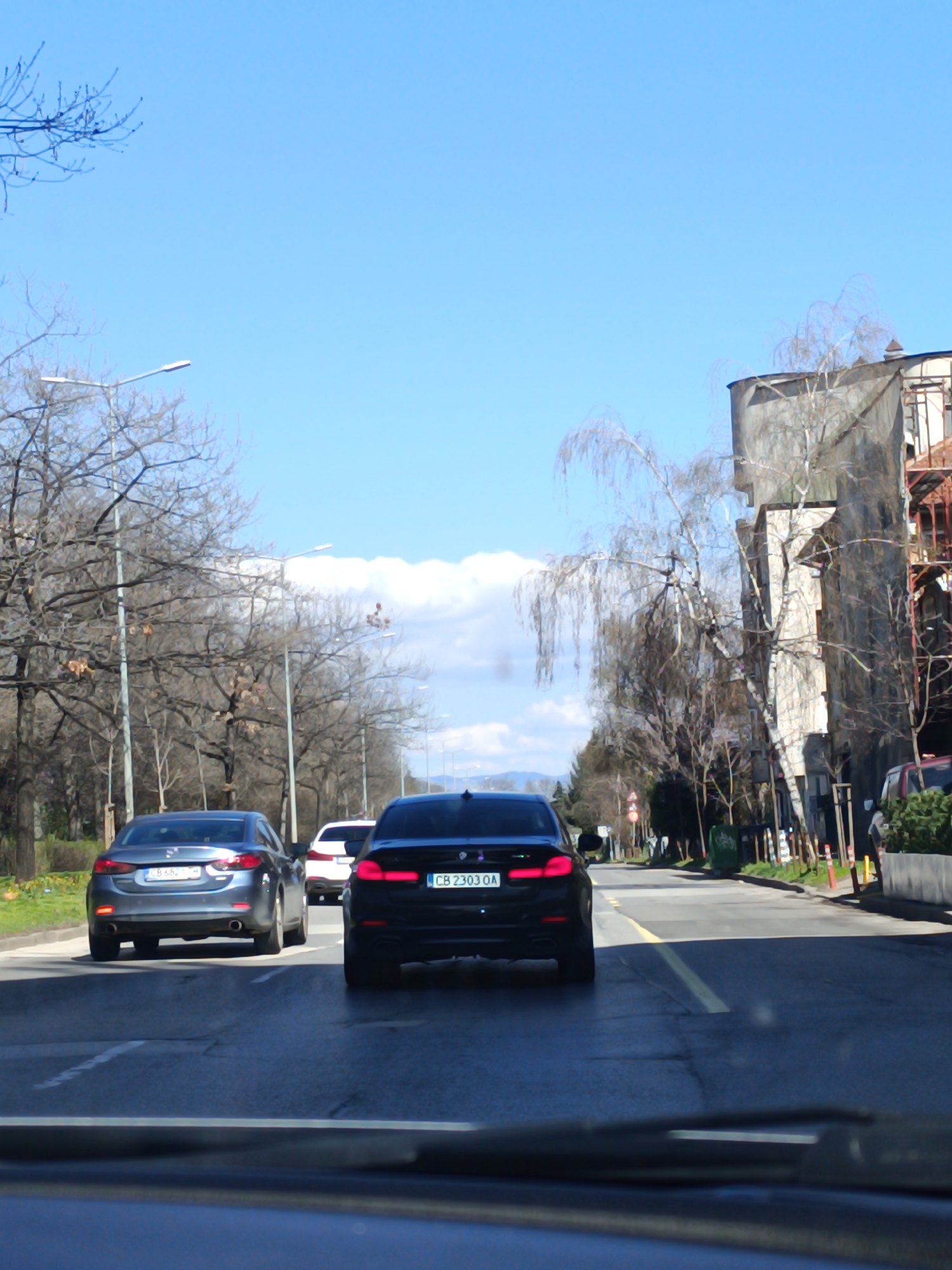 The image shows a view from inside a car driving on a road in Sofia, Bulgaria. The road is a two-lane road with a yellow line marking the center. There are two cars in front of the camera car: a black BMW with license plate "CB 2303 OA" and a gray Mazda with license plate "CB 8688 HM." There is also a white car in front of the black BMW. The cars are driving in the same direction. On either side of the road are trees and buildings. On the right side of the road is a tall building under construction with scaffolding. The sky is blue with some clouds. There are street lights on both sides of the road. Bare trees line the road. In the distance, mountains can be seen. The image is taken during the day.