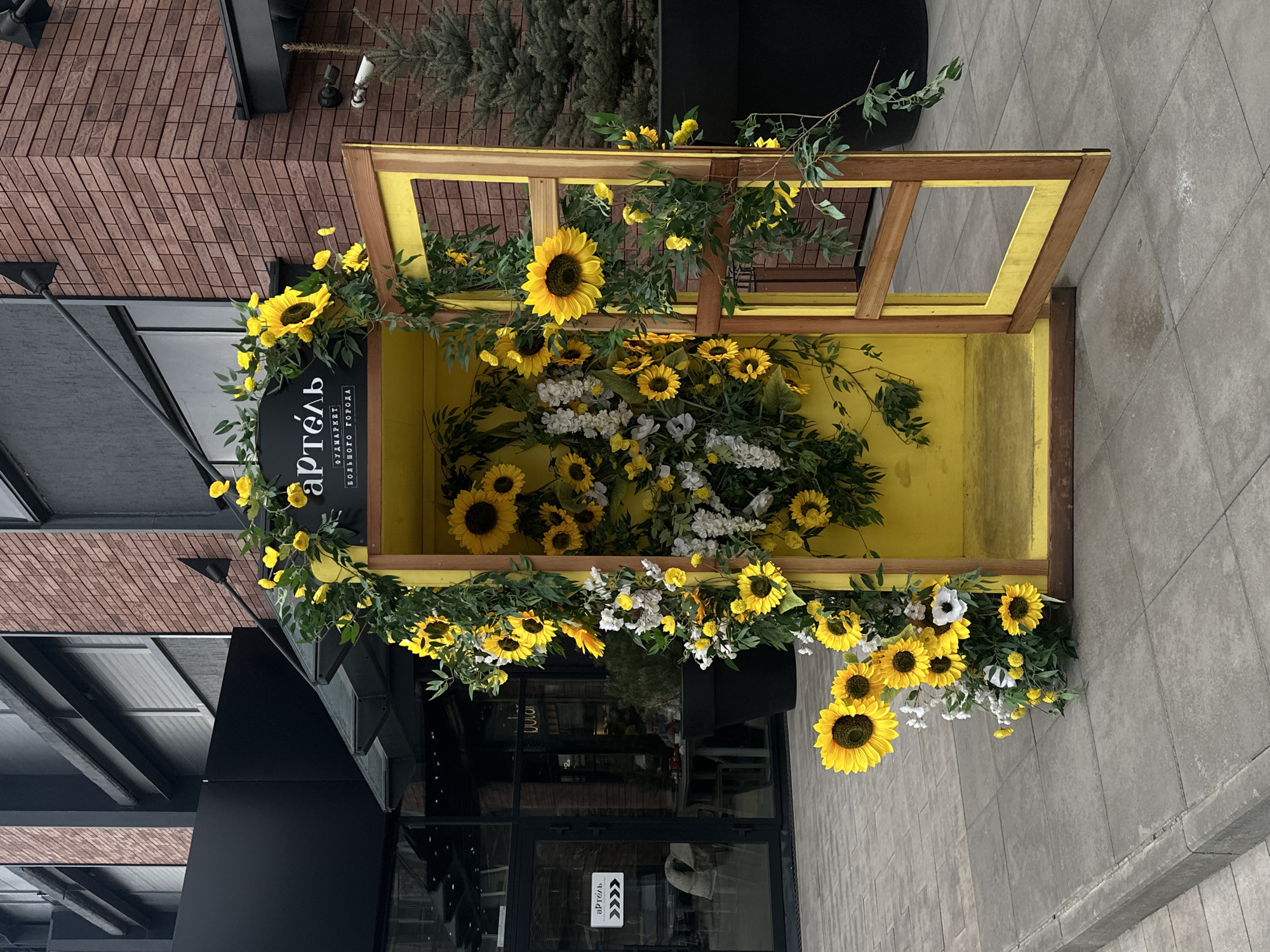 A partially overhead, ground-level outdoor shot captures a storefront display in Krasnoyarsk, Russia. The display is an open, yellow-painted wooden structure, decorated with sunflowers and white flowers. A dark sign labeled "артель. Фудмаркет Большого города," likely indicating a food market, is positioned on the left side of the wooden structure. The doorway of the building behind the display is glass with a small white sign. The exterior has a modern urban aesthetic with gray brick paving and a brick building in the background.