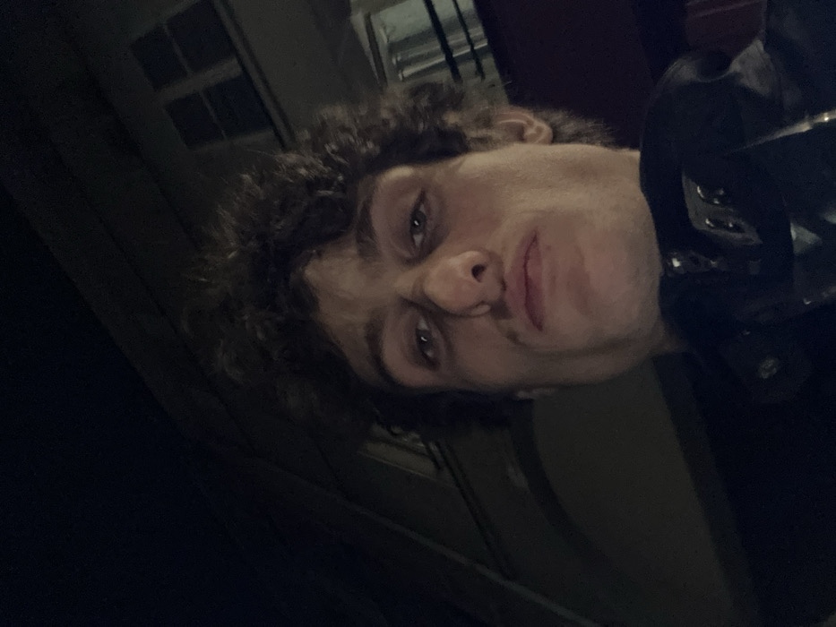 The image displays a close-up, low-angle self-portrait of a male subject with dark, tousled curly hair. The individual has a prominent nose, full lips, and a neutral expression, looking directly into the camera. He is wearing a dark-colored jacket, the collar and upper portion of which are visible in the lower frame. The background is dark, revealing architectural elements consistent with urban buildings. Specifically, parts of building facades are visible, including several dark rectangular windows on the upper right and a slight arch or curve on a lighter-toned structure in the upper left. These architectural characteristics are consistent with structures found in the city of Paris, France. The scene is dimly lit, suggesting nighttime or low-light conditions, with the subject's face illuminated by a frontal light source. No other individuals or specific objects are identifiable.