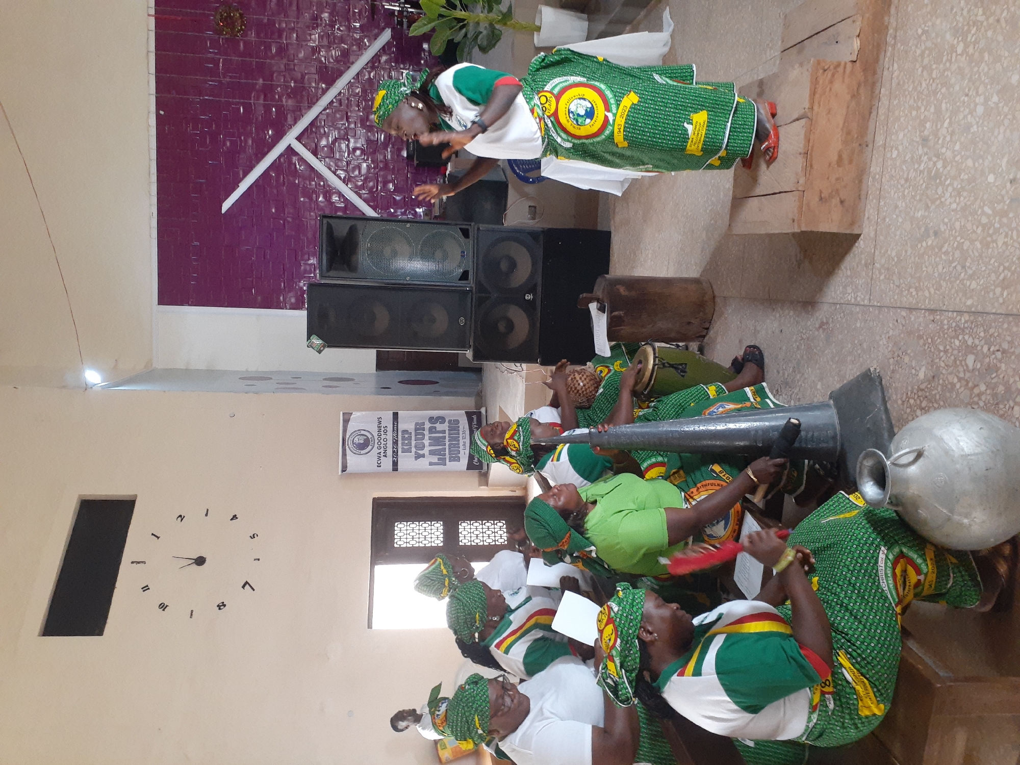 A brightly lit room in Barakin Lerawi, Nigeria, shows a gathering of people, predominantly women, dressed in green and white patterned traditional attire. One woman stands on a wooden platform near two tall speakers, gesturing animatedly as if addressing the group. In front of her, a row of women sits on the floor, some holding musical instruments, including what appears to be a large horn. A banner on the wall reads "ECWA GOODNEWS ANGLO-JOS" with the theme "KEEP YOUR LAMPS BURNING." A clock on the wall indicates the time is around 11:30. A decorative design is on the wall behind the speakers. A plant is visible near the standing woman. The room appears to be an event space, possibly a church or community hall.