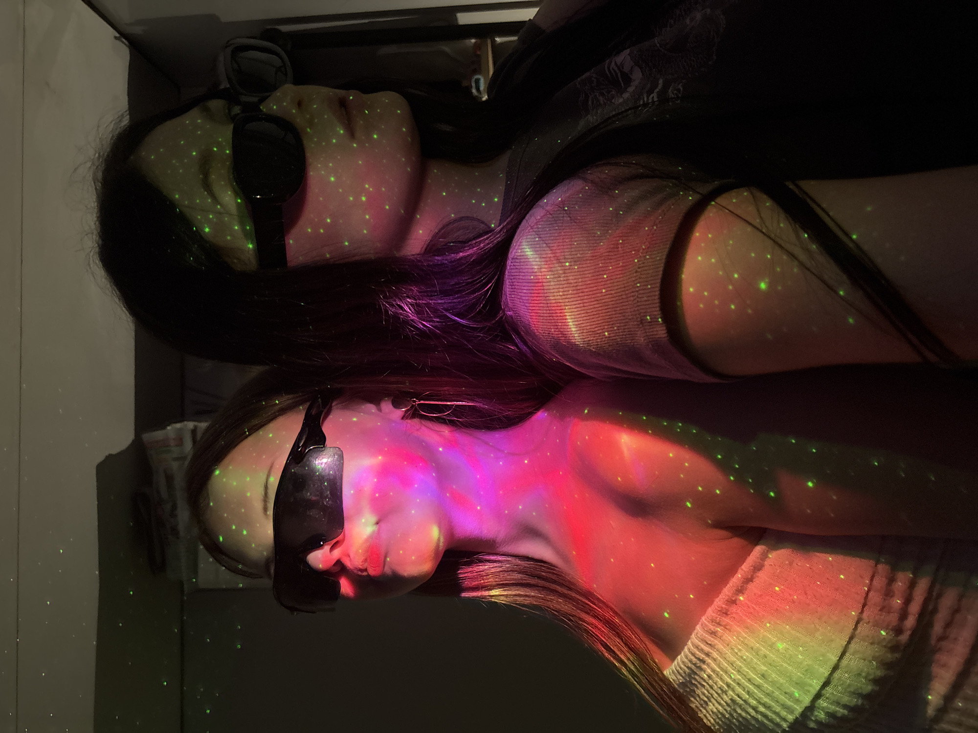 The image displays two young women in a dark indoor setting, illuminated by colorful light projections. Both subjects are positioned horizontally, appearing to be lying down or reclining, and are wearing dark sunglasses or eye masks that cover their eyes.
The woman in the foreground is seen in profile, with her face, neck, and shoulder prominently highlighted by a blend of red and purple light. Numerous small, bright green dots, resembling stars, are projected onto her skin, dark hair, and the light-colored, possibly knit, garment covering her shoulder.
Behind and slightly above her, a second woman is visible, also with dark hair and wearing similar dark eyewear. Her face and upper body are bathed in softer pink and green light, similarly adorned with the scattered green star-like dots. She appears to be wearing a darker top.
The background is indistinct and dark, indicative of a low-light environment. The overall scene suggests a relaxed atmosphere, possibly created by a mood-lighting projector that casts the speckled green lights and broad washes of red, purple, and pink hues across the subjects and surrounding surfaces. The women appear to be resting or enjoying the light display. There is no visible text in the image.