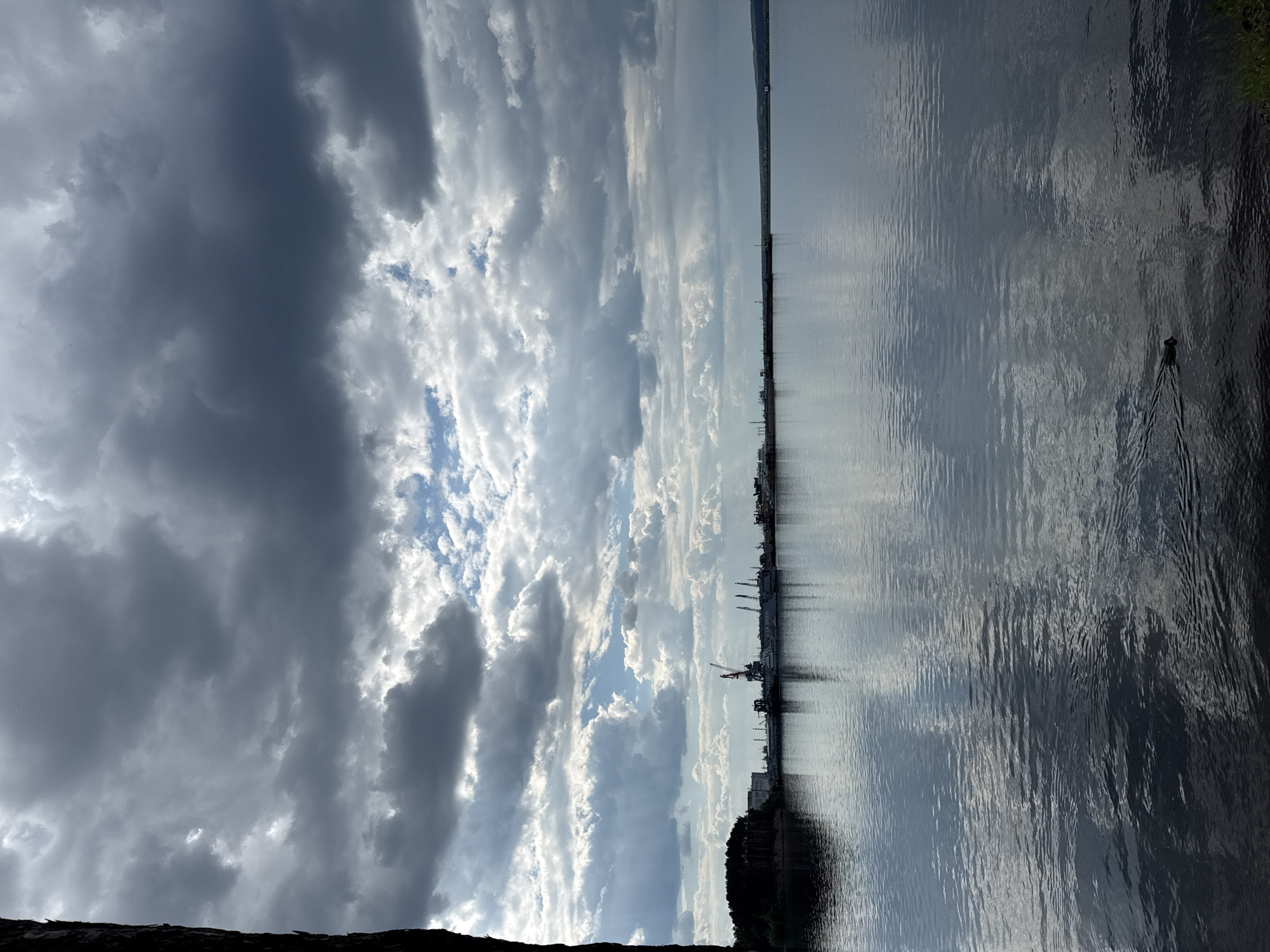 A dark, textured tree trunk is visible on the far left edge of the frame. The scene depicts a wide, calm body of water reflecting a dramatic sky filled with dark, heavy clouds interspersed with patches of bright blue and white. Along the distant horizon, a shoreline is visible, lined with trees on the left and industrial structures, including cranes and docked vessels, in the center. The water is smooth with ripples, and a single duck is swimming from right to left in the lower right quadrant, leaving a V-shaped wake. The overall atmosphere is overcast and somewhat moody. The scene is likely the Kama River in the city of Chaikovsky, Russia, given the presence of industrial port facilities along the river.