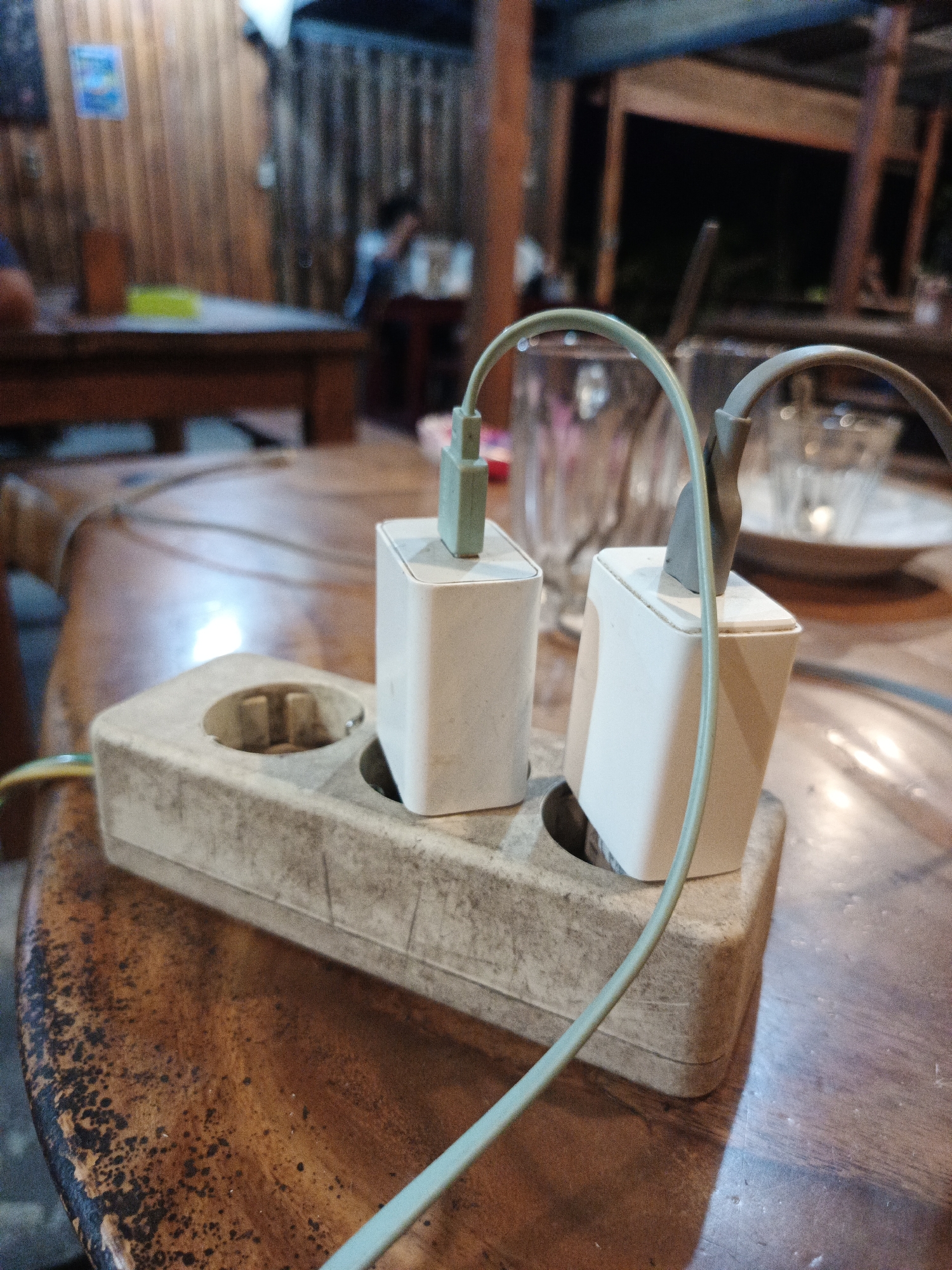 The image shows an electrical outlet on a wooden table in Geulumpangmeujim-Jim, Indonesia. Two white charging adapters are plugged into the outlet. Cables are connected to the adapters. In the background, there are tables and chairs with people sitting. Glasses and plates are visible on the tables. The environment appears to be an indoor space, possibly a cafe or restaurant.
