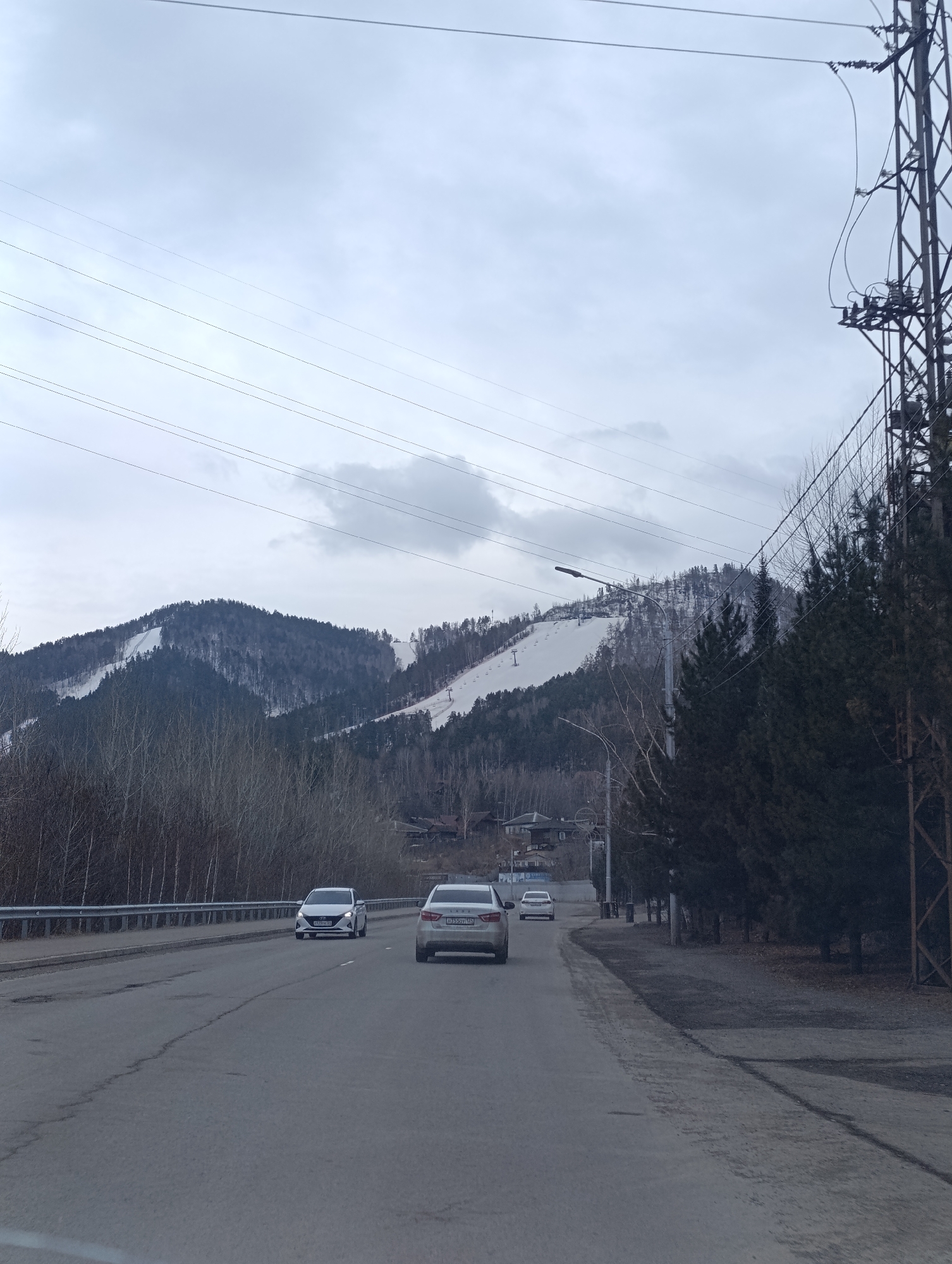 The image depicts a road leading towards a snow-covered mountain with visible ski slopes. Three cars are present on the road, with one in the foreground and two further away. Trees line the right side of the road, and a metal barrier is visible on the left. Overhead power lines crisscross the sky. The location is Krasnoyarsk, Russia.