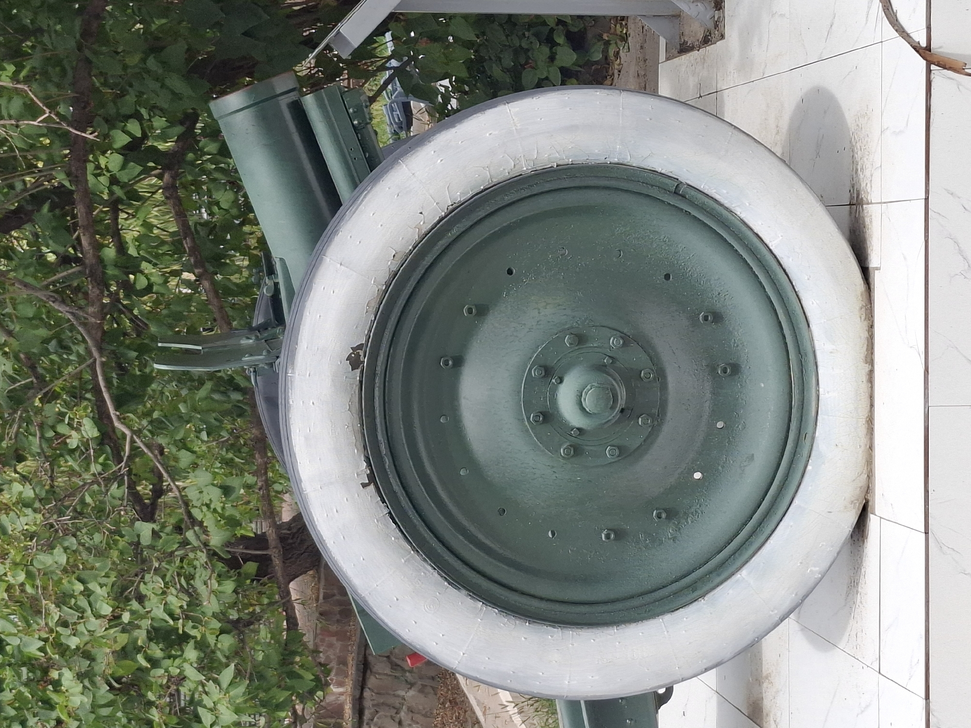 The image shows a close-up view of a large, old, green-painted wheel, likely from a piece of artillery or military equipment. The wheel has a light-colored, worn tire with visible signs of cracking and peeling. The rim of the wheel is also green and features several bolts. The background consists of green foliage from a tree and some stone and metal structures, possibly part of a monument or display. The ground beneath the wheel is tiled. There are no people visible in the image. The object appears to be stationary and displayed outdoors.