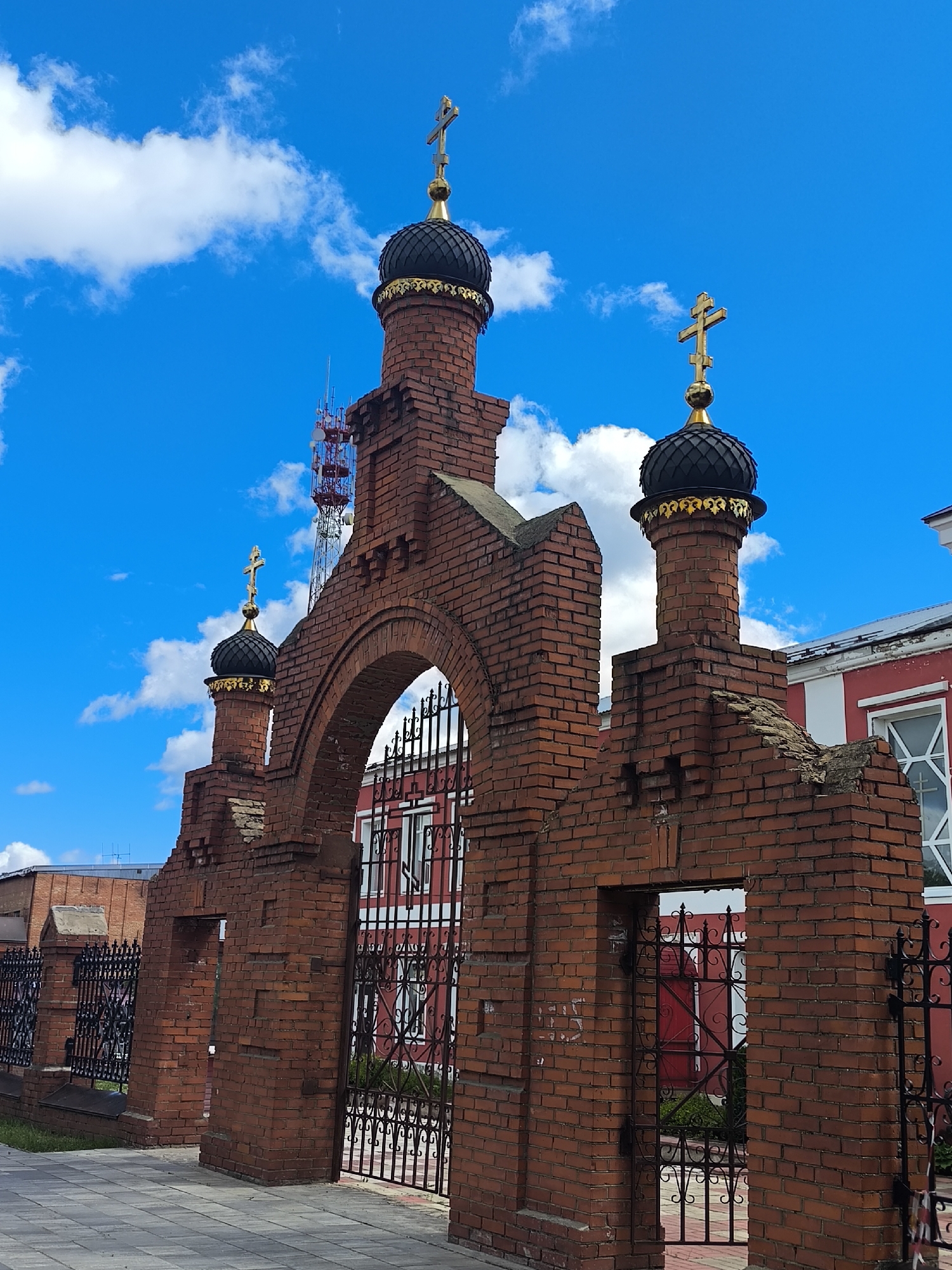 This is a frontal, low-angle shot of a red brick gate structure in Taldom, Russia, against a bright blue sky with scattered white clouds. The gate features multiple arched openings and brick towers topped with black domes and gold crosses. In the background, a red building with white trim and windows is visible, as is a black wrought-iron fence. A cellular tower with red and white sections stands behind the gate. The ground in front of the gate is paved with gray square tiles.