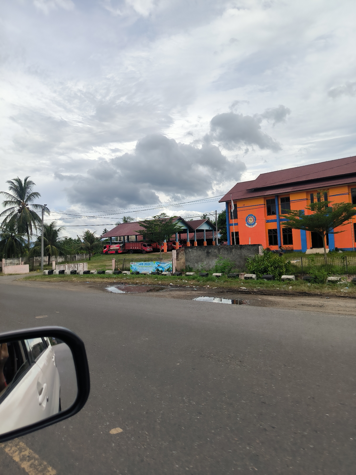 This media file captures a roadside view from a moving vehicle in Reuhattuha, Indonesia, on an overcast day. The lower-left corner prominently features a car's side mirror, reflecting part of a white vehicle. The asphalt road in the foreground and mid-ground is marked with some cracks and small puddles, suggesting recent rainfall.
On the right side of the road, behind a low concrete barrier and some unkempt greenery, stands a vibrant two-story building. It features a reddish-brown tiled roof and is painted bright orange with distinctive blue trim around its windows and corners. A circular emblem is visible on its front wall, and a white flag flies from an adjacent pole. To its left, a single-story structure with a similar roof appears, painted in red and green/blue. Parked near this building are several red trucks, one of which clearly displays "BPBD" (Badan Penanggulangan Bencana Daerah - Regional Disaster Management Agency) on its side. Further in the background, more buildings with similar architectural styles are interspersed among dense tropical vegetation.
The left side of the road is lined with several tall palm trees and other lush tropical foliage. The sky is predominantly covered by thick, grey clouds, indicating an overcast and potentially humid atmosphere. A blue banner or billboard is visible near the red and green building, displaying the text "HKB 2026," "Amanah Untuk Indonesia Maju," and the "BPBD" logo, suggesting a disaster preparedness event or campaign. The scene depicts a neutral streetscape, without any specific human activity, under daytime conditions.