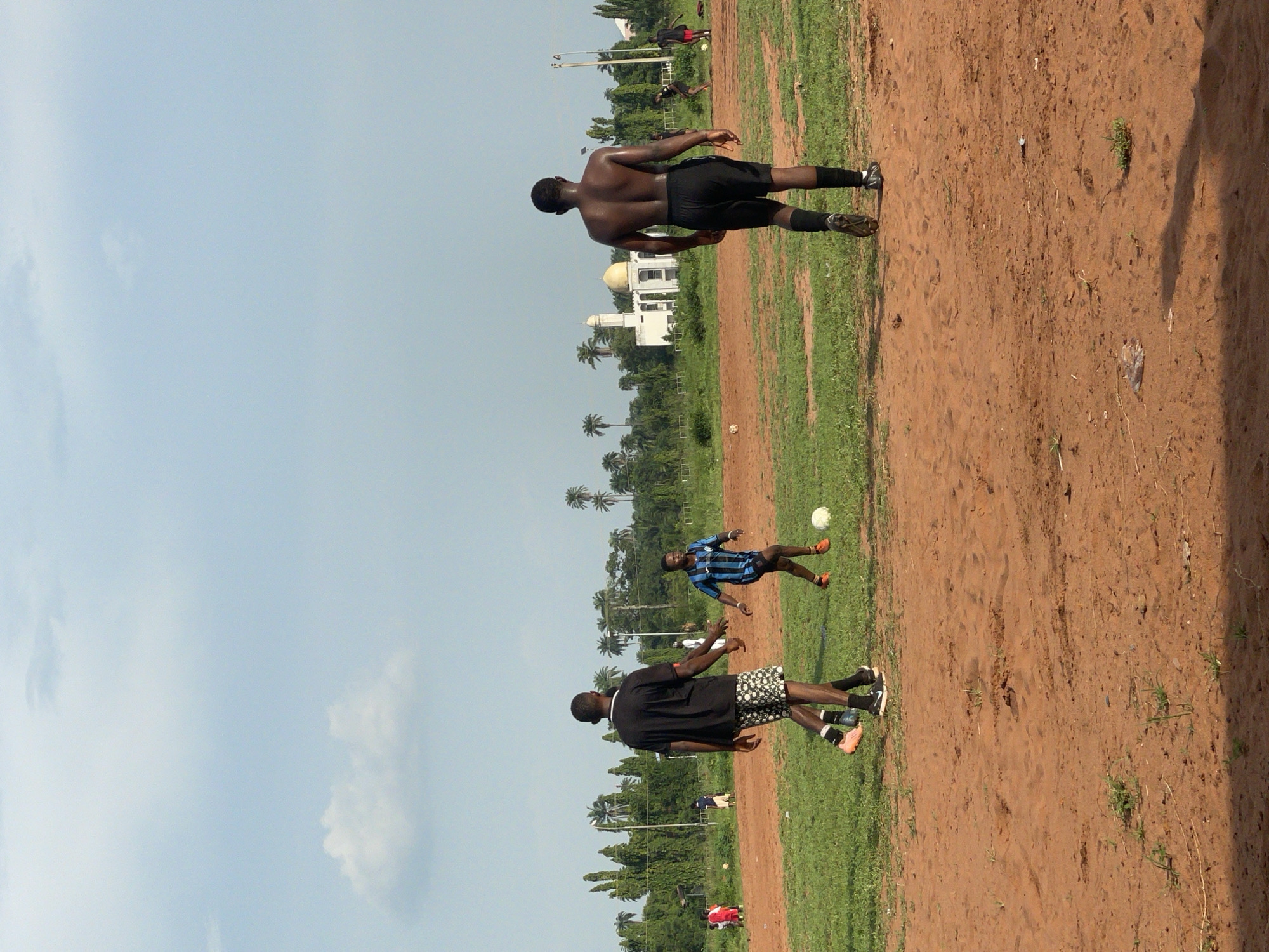 This image, taken in Ogbogbo, Nigeria, captures an outdoor soccer (football) game in progress. The scene is set on a field characterized by patches of green grass mixed with reddish-brown dirt and sand under a bright, light blue sky with scattered clouds.
In the foreground, two men are seen from behind. One man, shirtless and wearing black athletic shorts and black socks, stands with his back to the camera, his skin appearing glossy from sweat. Beside him, another player wears a black t-shirt and patterned black and white shorts, also facing away from the viewer, observing the game.
Further into the field, a third player, dressed in a blue and black striped jersey and black shorts, is actively engaged with a white soccer ball, appearing to kick it. Several other figures are visible in the mid-ground and background, participating in or watching the game.
The background features a prominent white building topped with a golden dome, resembling a mosque, partially obscured by lush green trees, including several tall palm trees. The clear sky and bright light suggest it is daytime, likely during sunny weather. No discernible text is visible within the image. The overall impression is a vibrant, casual soccer match taking place in a warm, tropical setting.