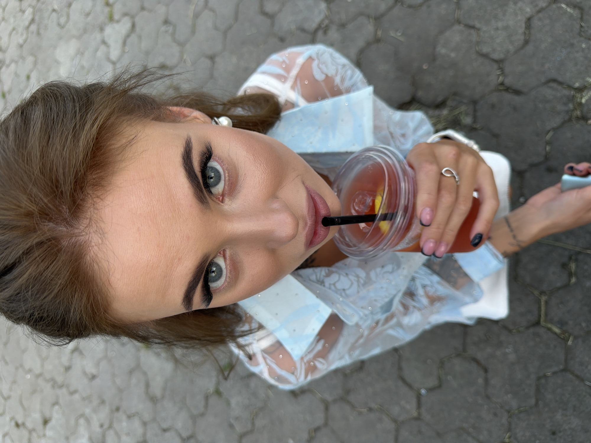In this overhead shot, a young woman with light blue eyes and dark eyeliner is captured from a high angle. She has light brown hair that is slightly messy and styled with a pearl earring on her left ear. She is holding a clear plastic cup filled with a reddish-orange drink that contains ice and fruit, with a black straw inserted. The woman is wearing a sheer white garment with a light blue collar, adorned with a subtle lace pattern. She is also wearing a silver ring on her right hand and has black and pink manicured fingernails. Her lips are slightly parted as she appears to be drinking from the cup. The background consists of a grey, hexagonal paver stone ground, suggesting an outdoor setting. The lighting suggests it is daytime. The overall impression is a candid moment of someone enjoying a refreshing drink outdoors.