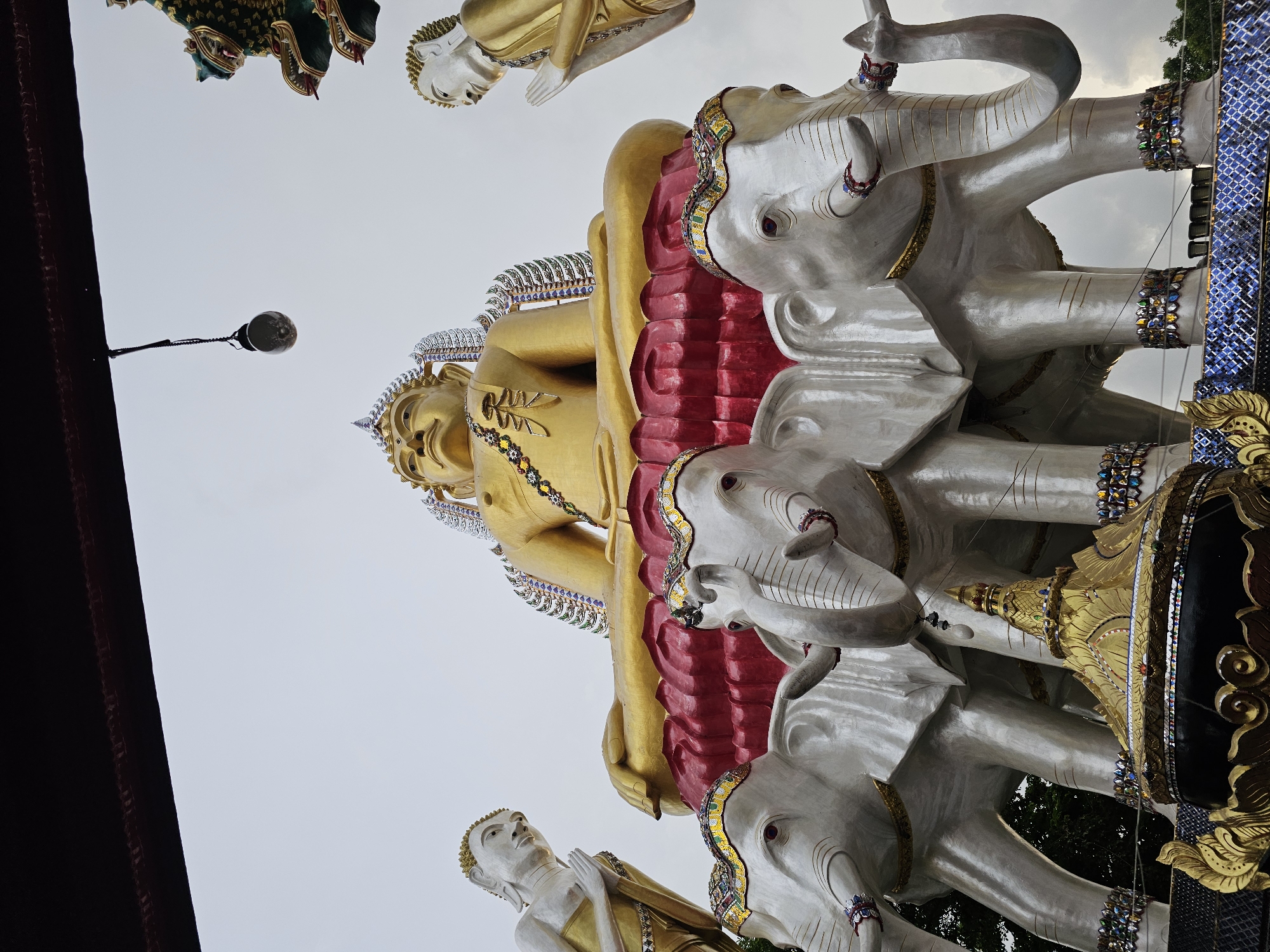 This is a low-angle, full-frame, eye-level photograph of a large Buddhist statue, likely located in Bangkok, Thailand, given the stylistic elements. The central subject is a golden statue of the Buddha in a seated meditation pose, adorned with colorful, jeweled ornaments around its neck and on its elaborate headdress. The Buddha sits atop a vibrant red and gold lotus-like structure.
Flanking the central Buddha statue are two smaller, silver statues of Buddhist figures, both with hands pressed together in a gesture of prayer or respect. These figures also wear golden robes.
Supporting the entire central structure are three large, white statues of elephants. These elephants are also adorned with colorful, jeweled decorations on their foreheads, tusks, and legs, and they appear to be walking in unison. In front of the elephants, a gold and black ornate shrine-like structure is visible.
The background consists of a muted, overcast sky. In the upper portion of the image, a dark, possibly wooden, architectural element overhangs the scene. A single light bulb hangs from a wire, casting a faint glow. On the far right, a glimpse of a decorative green dragon or mythical creature, likely part of the temple's architecture, can be seen. The overall atmosphere is one of solemnity and grandeur, typical of religious sites.