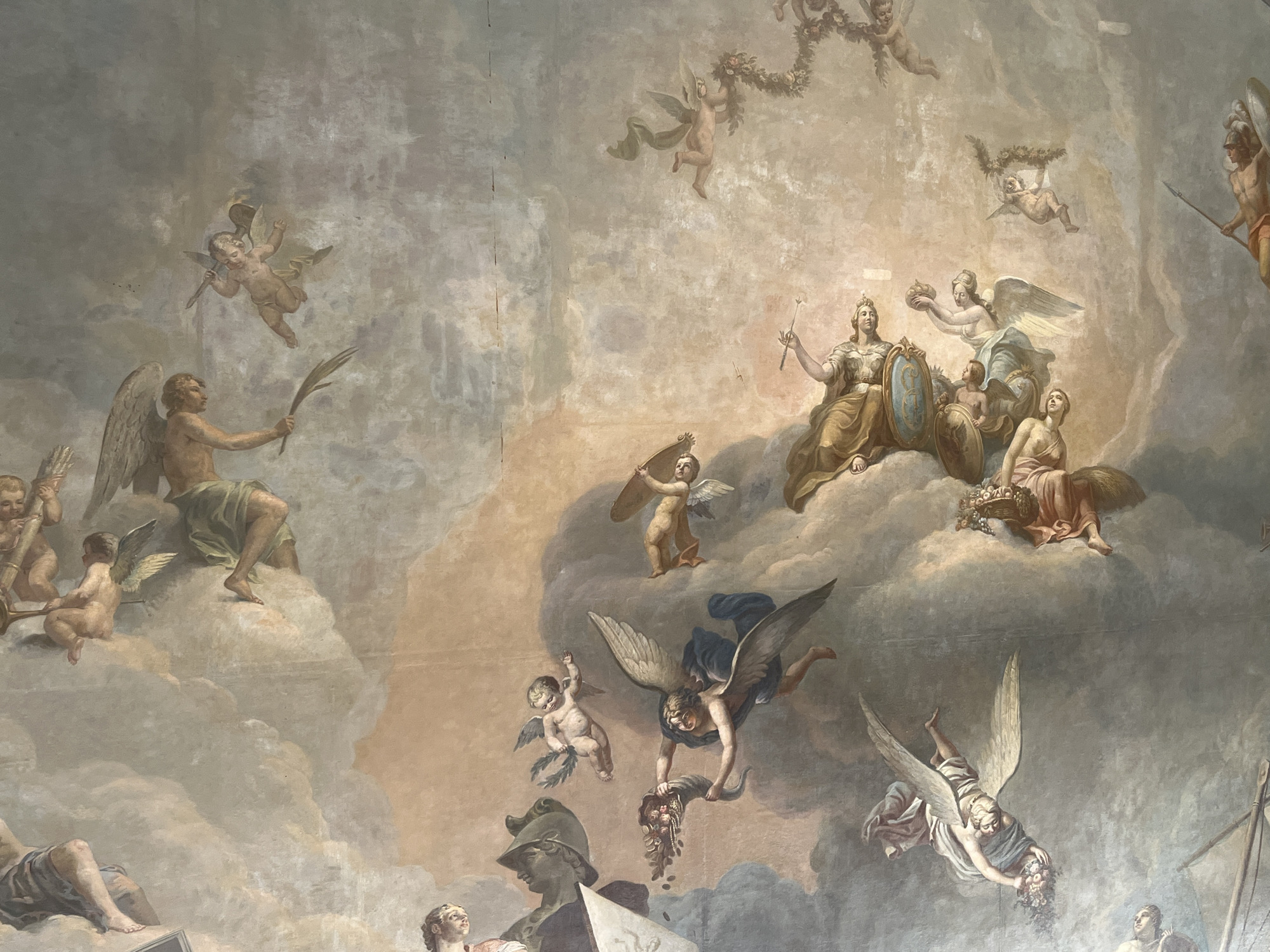The image depicts a section of a fresco, likely a ceiling painting, featuring various mythological figures and cherubic angels amidst a swirling cloudscape. Central figures include a regal, crowned woman holding a spear and shield, potentially an allegory of wisdom or justice, surrounded by attendants and angels bearing wreaths and crowns. Other figures present include partially draped men and winged cherubs, some carrying musical instruments or symbolic objects. The palette is dominated by muted tones of cream, ochre, gray, and hints of blue, characteristic of classical or Renaissance-era paintings. The location is unknown.