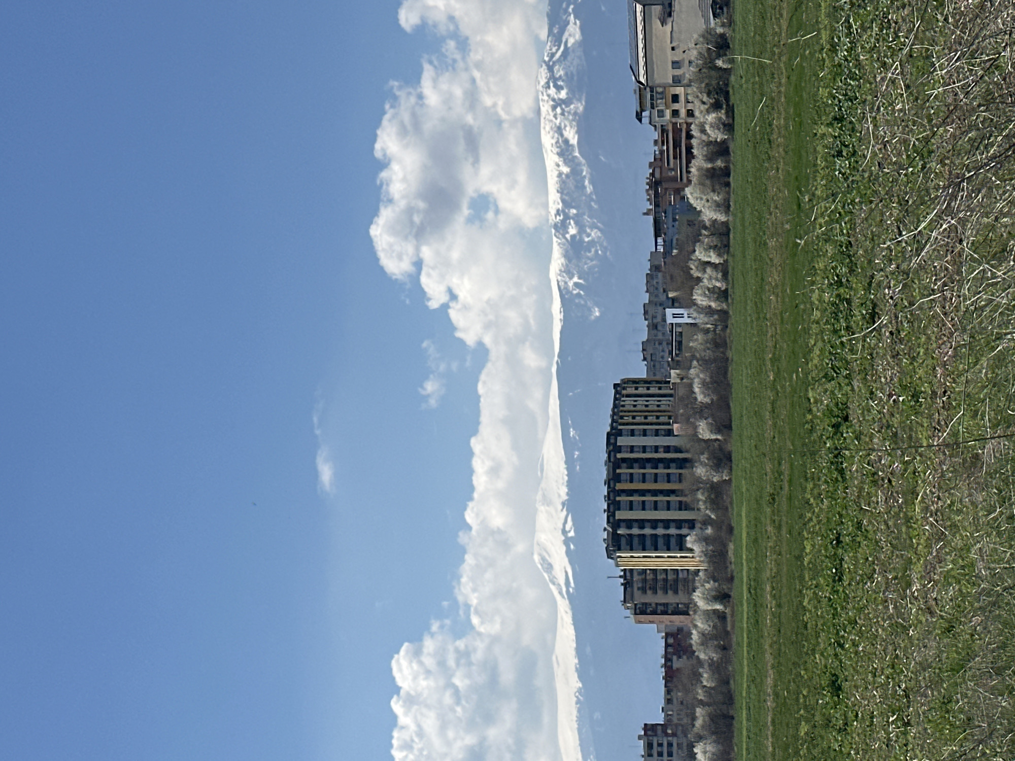 The image shows a view in Sofia, Bulgaria, with a clear blue sky and white clouds to the left. A line of buildings is seen to the right of the clouds. A long green field occupies the lower right portion of the image, transitioning to a shrubby area on the far right. In the background, a mountain range is partially visible, blending into the cloud line. The image has been rotated.