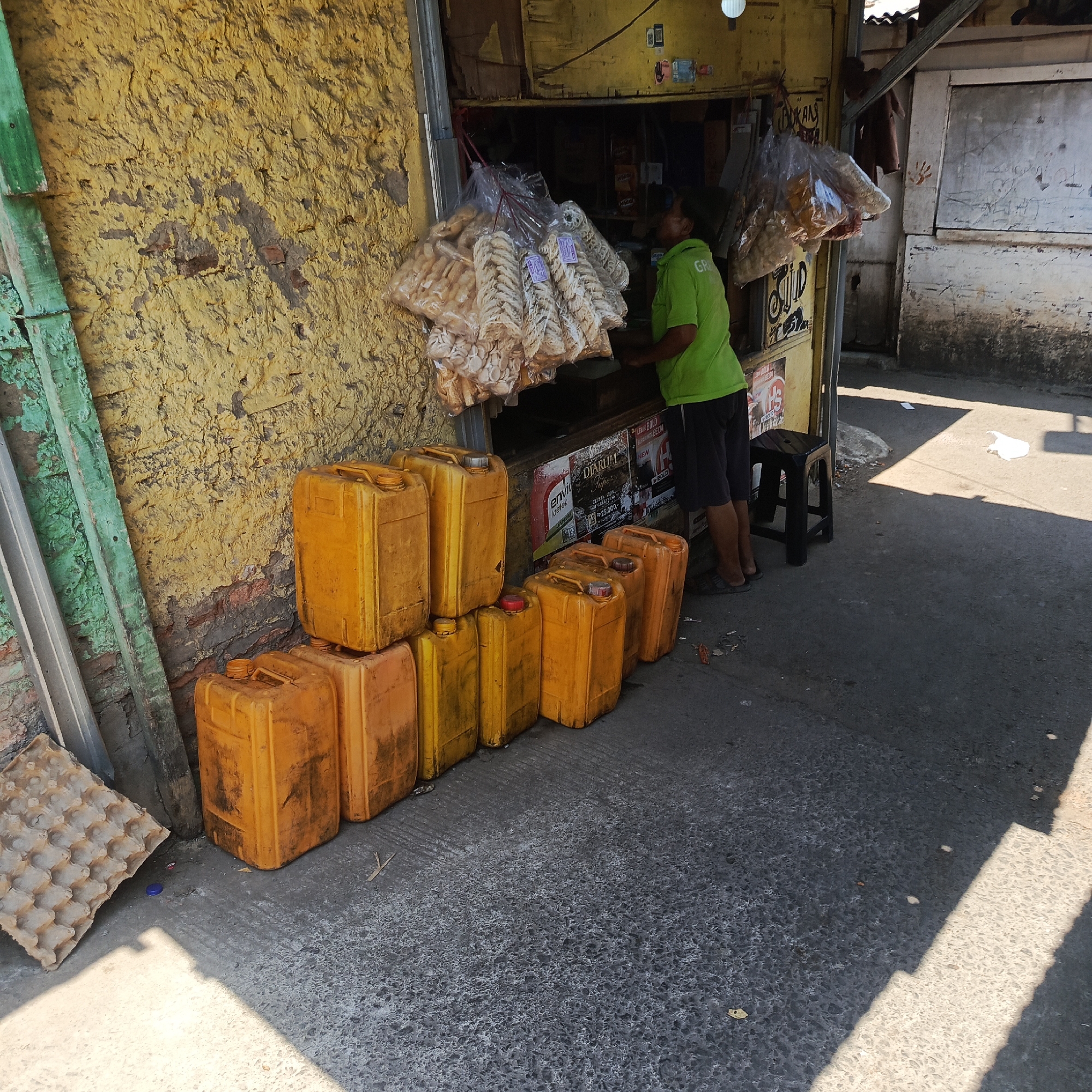 The image shows an outdoor scene in South Jakarta, Indonesia, likely on a street or alleyway adjacent to a small shop. In the foreground, a stack of large, orange-yellow plastic jerrycans is arranged on the ground. To the left, an egg carton made of corrugated cardboard is propped against a rough, yellow-painted wall. A person wearing a bright green t-shirt and dark shorts stands behind a counter inside a small shop or stall. The stall is partially visible and appears to be selling packaged snacks that are hanging from the ceiling in plastic bags. The wall of the shop is weathered and cracked. Sunlight casts strong shadows across the scene, indicating it is daytime. The ground is paved with asphalt and shows some debris.