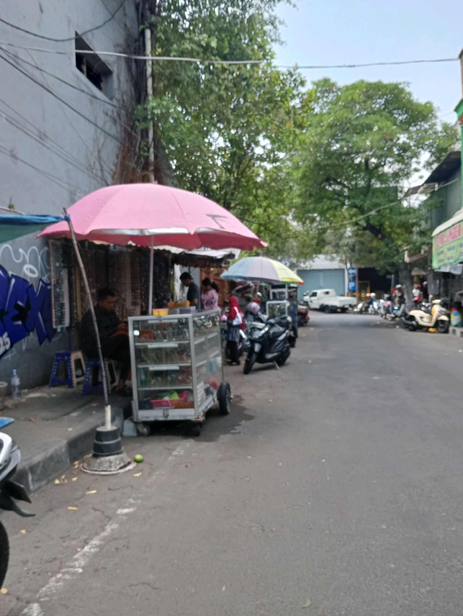 A street scene in Jagalan, Indonesia shows a row of shops and stalls lining a paved road. In the foreground, a pink umbrella shades a display cart filled with various items, including what appear to be sunglasses and other accessories. A person sits behind the cart. To the left, a blue stool is visible next to a graffiti-covered wall. Further down the road, several motorcycles are parked, and more stalls and shops are seen, some with awnings. A white pickup truck is parked further in the distance. The sky is overcast, and trees are visible in the background. The overall impression is that of a busy marketplace or commercial street.