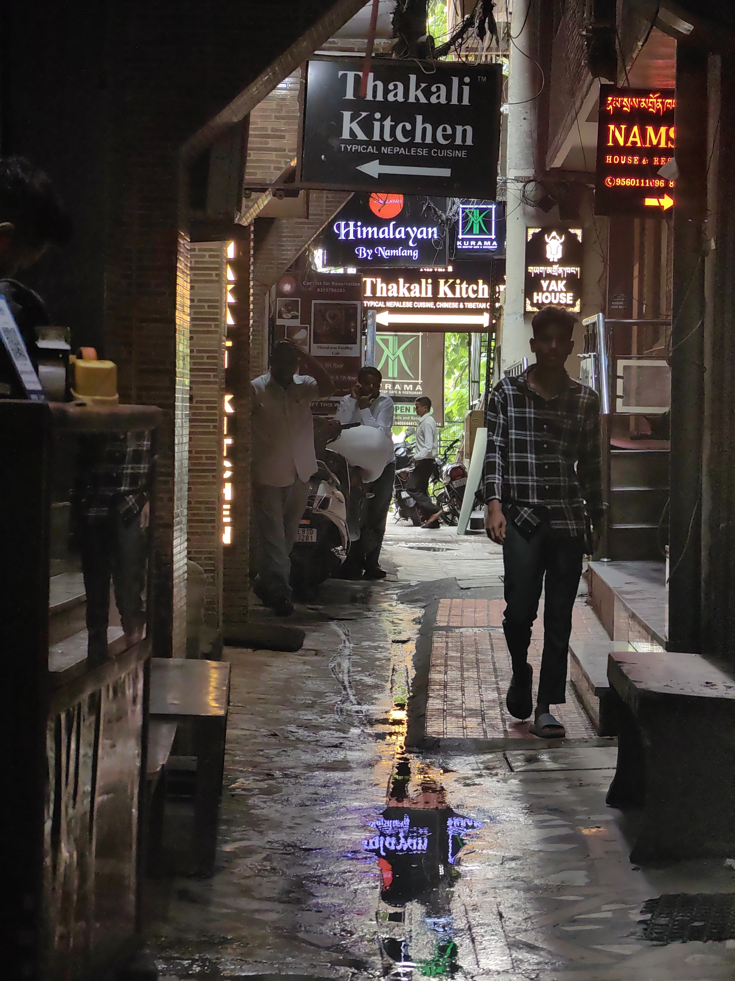 The image depicts a narrow alleyway in Delhi, India, likely in a market or commercial area, characterized by a wet, reflective ground surface suggesting recent rain. Several signs for restaurants are visible, including "Thakali Kitchen" with the tagline "TYPICAL NEPALI CUISINE," "Himalayan by Namlang," "KURAMA RESTOP CAFE & RESTAURANT," and "YAK HOUSE." Another sign, partially obscured, reads "NAMS HOUSE & RESTAURANT."
In the foreground, a young man wearing a plaid shirt and dark trousers walks away from the camera. Further down the alley, two men are engaged in conversation near a parked motorcycle. One man, in a white shirt and grey trousers, is gesturing with his hand. Another man, in a white shirt, is seated. The lighting is dim, with neon signs and overhead lights casting reflections on the wet pavement. The architecture suggests a dense urban environment with brick and concrete structures.