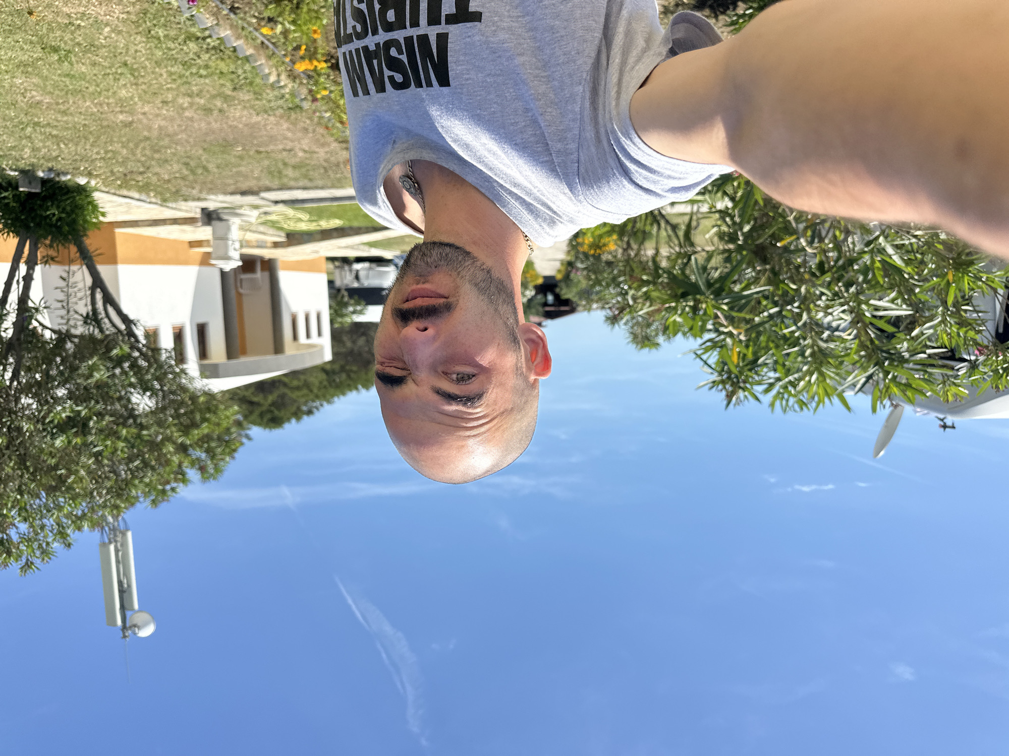 A selfie of a bald man with a short beard, wearing a grey t-shirt with "NISAN TURIST" printed on it and a silver chain, winking at the camera. The photo appears to have been taken outdoors in Donji Štoj, Montenegro on a sunny day, with a clear blue sky visible. In the background, there is a building with white and orange exterior, green grass, trees, a light-colored building, a dark car and an antenna.