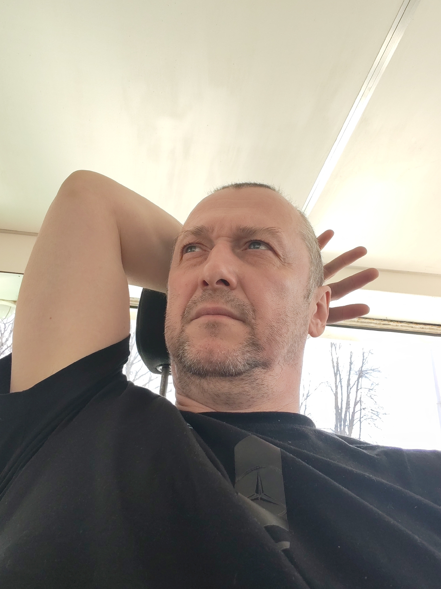 A medium close-up shot shows a middle-aged Caucasian man with short, balding hair and stubble. He's wearing a black t-shirt with a logo that appears to be a Mercedes-Benz emblem. His right arm is raised and bent, with his hand placed behind his head. The background is a bright, neutral color, likely the ceiling of a room. A window is visible in the upper right corner, showing bare trees outside. The headrest of a car seat can be seen behind his right arm. There is no information to confirm that the location is in the city of Yakhroma, Russia.