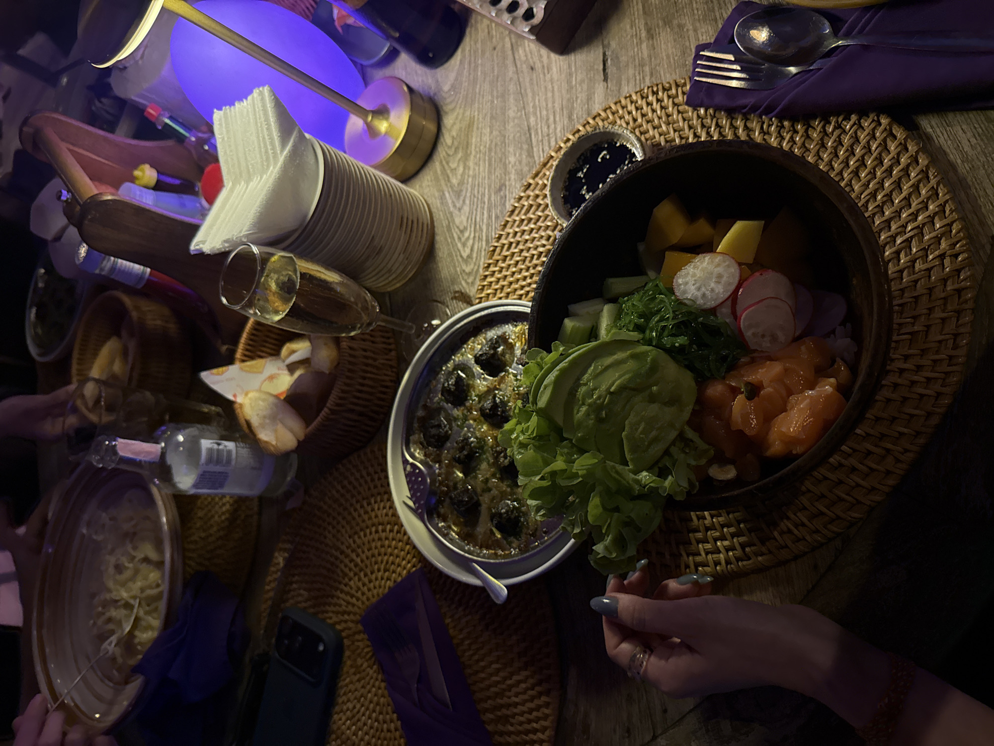 This detailed description of the scene in the city of บ้านบางรักษ์, Thailand, shows an indoor dining table illuminated by dim, warm ambient light, with a prominent purple decorative light in the background. **People:** At least two individuals are present, indicated by visible hands. One person's left hand, with manicured grey/blue nails and two rings, is positioned in the lower-left foreground near a poke bowl. Another person's hand, holding a fork, is visible in the upper-left, reaching into a large pasta bowl. **Food & Beverages:** * **Pasta:** A large, round, light-colored ceramic bowl in the upper-left contains a substantial serving of pasta with a light-colored sauce, into which a fork is inserted. * **Escargot/Mushroom Dish:** A white round plate in the center-left holds a dark metal or ceramic dish filled with multiple dark, round items, possibly escargots or baked mushrooms, submerged in a glistening, herbaceous sauce. A small, two-tined fork is placed inside the dish. * **Poke Bowl:** A dark brown wooden bowl in the center-right foreground features a colorful poke bowl. Visible ingredients include diced raw salmon, sliced avocado, green leafy lettuce, seaweed salad, cubed mango, and sliced radishes. * **Dipping Sauce:** A small white ceramic bowl adjacent to the poke bowl contains a dark liquid, likely soy sauce, with sesame seeds. * **Bread:** A small woven basket in the upper-center holds several pieces of white bread. * **Drinks:** A tall, slender champagne flute in the center-left contains a light golden sparkling beverage. A clear glass bottle with a white label and pink foil neck, possibly sparkling water, stands next to the pasta bowl. **Tableware & Decor:** * **Table Surface:** The table is constructed of dark wooden planks. * **Placemats:** Several round, woven straw or rattan placemats are underneath the dishes. * **Napkins:** Purple fabric napkins are visible, one neatly folded with cutlery on the right, and another near a smartphone on the left. A stack of white paper napkins is in a white holder in the center-right. * **Cutlery:** Stainless steel forks and spoons are present, some wrapped in napkins. * **Smartphone:** A dark-colored smartphone encased in a protective cover rests on a placemat in the lower-left. * **Lighting:** A small, modern, brass-colored table lamp with a dark shade provides focused illumination on the right side of the table. A large, glowing purple spherical or egg-shaped decorative light is situated behind the table lamp. * **Condiments:** A wooden caddy