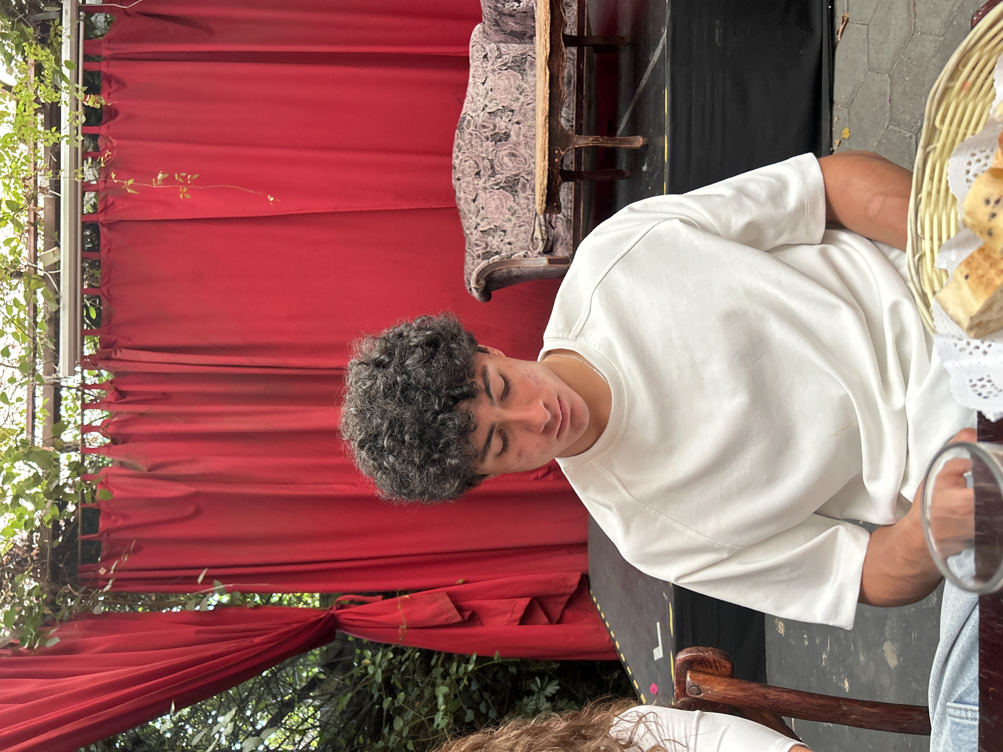A medium shot shows a young man seated at a round table, partially obscured by a wicker basket containing food items, and a glass. He is wearing a white long-sleeved shirt and appears to be looking down. The setting includes a backdrop of red drapes and greenery, along with a vintage-style sofa partially visible in the background. The location appears to be an outdoor dining area. The city and location are unknown.