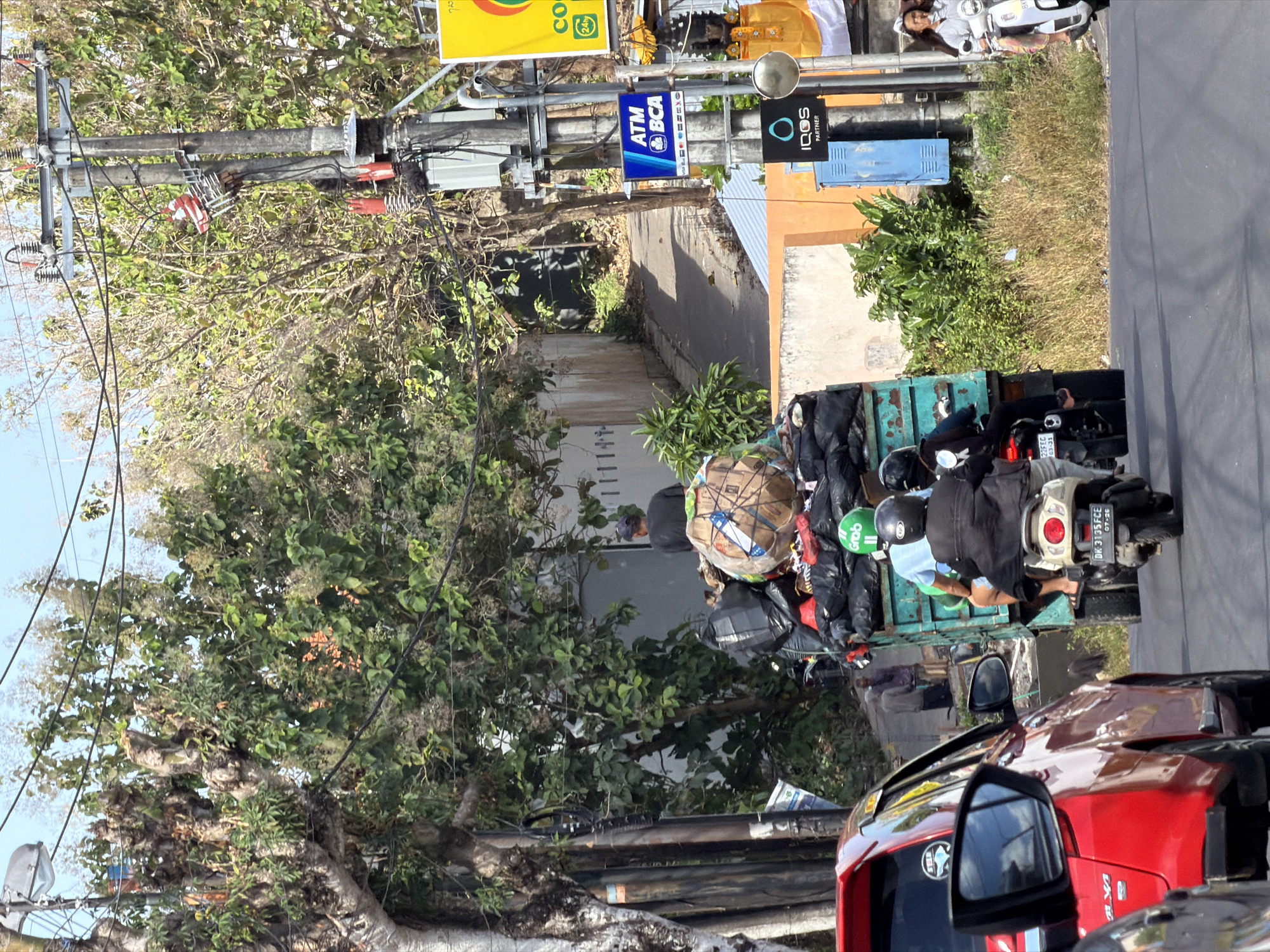 The image depicts a street scene in Kuta Selatan, Indonesia, with a focus on traffic and urban elements. In the foreground, a red car is visible, partially obscuring a motorcycle carrying a rider. Further down the road, a green truck is overloaded with trash bags and large circular objects wrapped in brown paper. Two individuals are seated on the truck, with one wearing a green helmet and the other a black helmet. The truck is moving in the same direction as the vehicles in the foreground. On the right side of the road, there is a utility pole with various signs and electrical equipment. One sign reads "ATM BCA," indicating a banking service. Another sign displays the logo for "IQOS PARTNER." A woman on a white scooter is also visible on the right side of the road, riding away from the camera. The background shows lush green trees and vegetation, suggesting a tropical environment. The overall scene conveys a sense of everyday life and commerce in a busy Indonesian city.