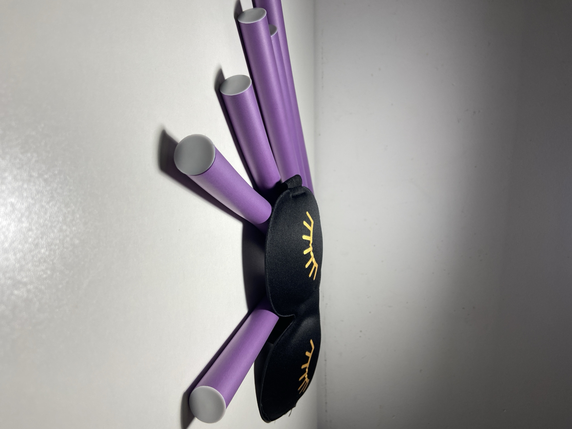 The image depicts a black sleep mask adorned with gold eyelash designs. The mask is draped over several purple rods with gray tips, which are positioned against a white wall. The setting appears to be indoors, with light and shadow play suggesting an artificial light source. There are no people visible. The location in the city of Budva, Montenegro is not deducible from the image.