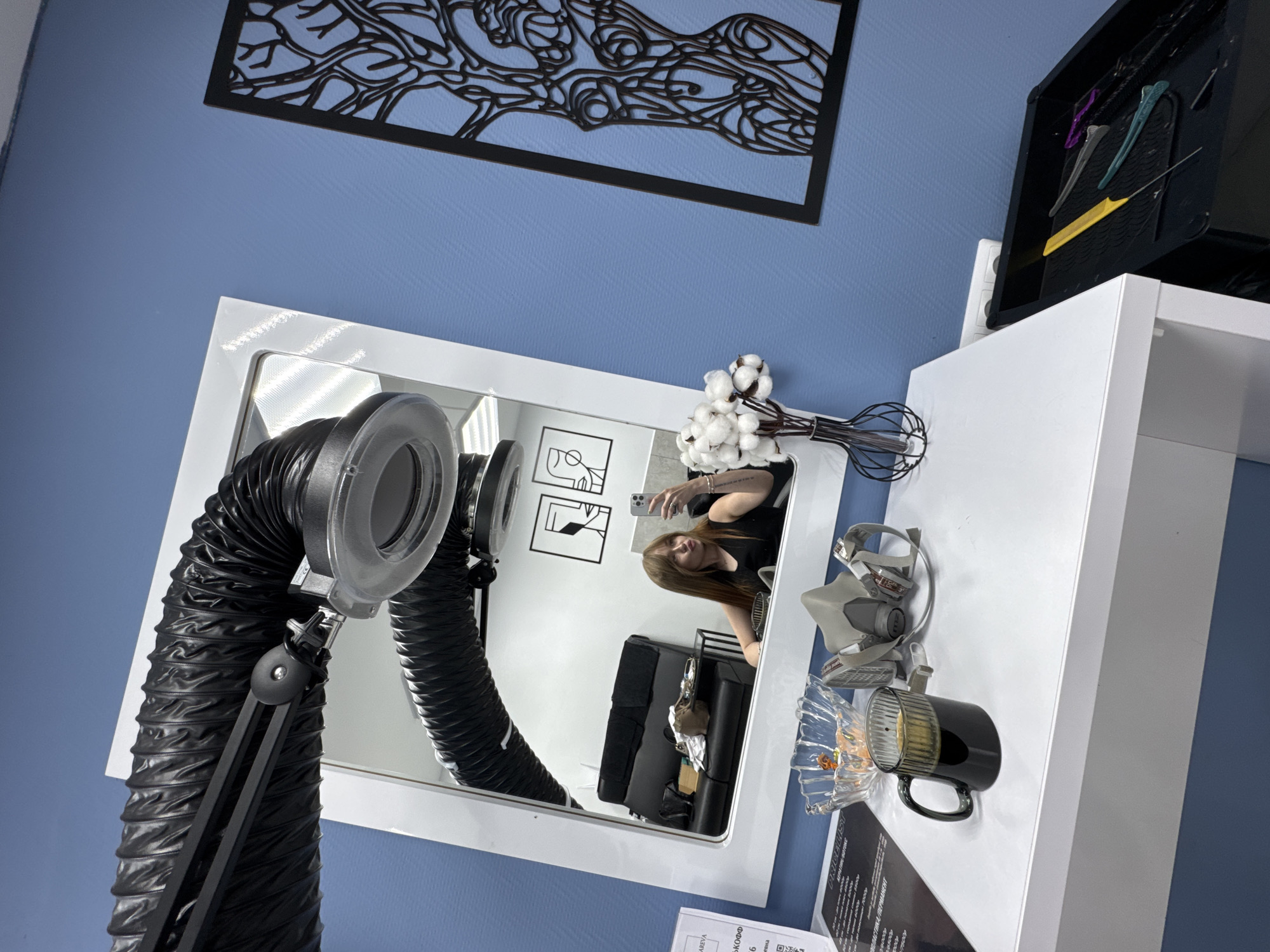 A young woman with long, light brown hair is taking a mirror selfie in what appears to be a salon or treatment room. She is wearing a black sleeveless top and is seated, looking towards her phone. Reflected in the large, white-framed mirror behind her, a black leather couch with some items on it, and modern art with geometric figures can be seen. In the foreground, on a white surface, there is a clear glass bowl with orange objects, a gray respirator mask with straps, and a dark brown mug filled with a dark liquid. To the right of the mug, a thin-stemmed vase holding white cotton-like flowers is placed. A black exhaust hose with a flexible ribbed texture extends from the top left corner of the frame, connected to a light fixture above the mirror. The wall behind the table and mirror is painted a light blue, with a black, stylized, abstract artwork featuring a branching pattern resembling a tree or body outline on the right side. To the far right, a black tray holds various hair styling tools including combs and clips. The overall impression is a personal space within a professional setting, possibly during a break or after a procedure. The image does not provide enough visual information to determine the specific location within the city of Chaikovsky, Russia.