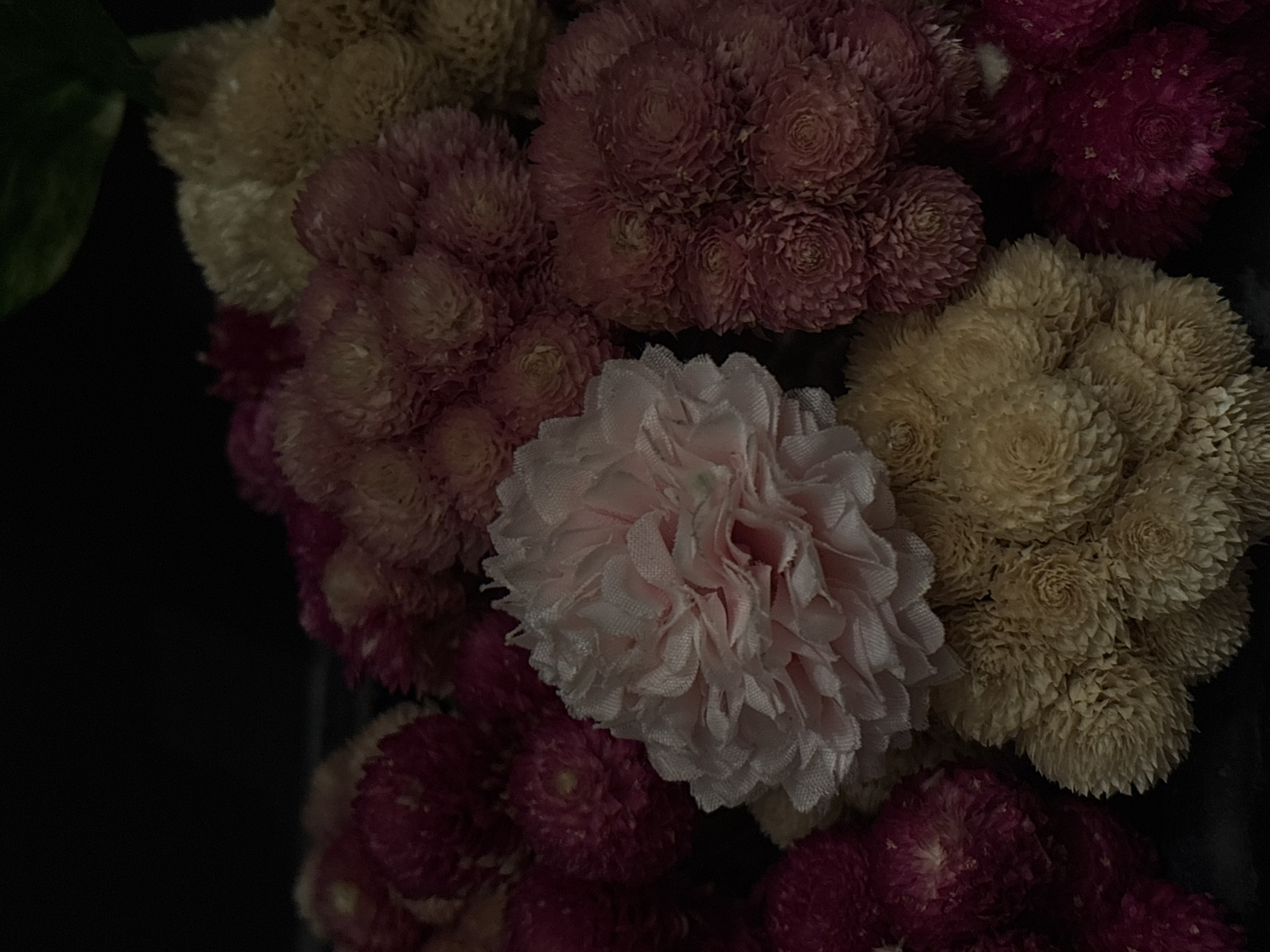 A close-up photograph shows a cluster of dried flowers in various colors, including pale yellow, pink, and dark red. The arrangement is dense and tightly packed, with some flowers exhibiting a spherical shape with intricate petal arrangements. A larger, lighter pink flower with more delicate petals is centrally positioned among the tighter, darker blooms. The background is a dark, indistinct space, highlighting the colors and textures of the flowers. Location and date are not determined.