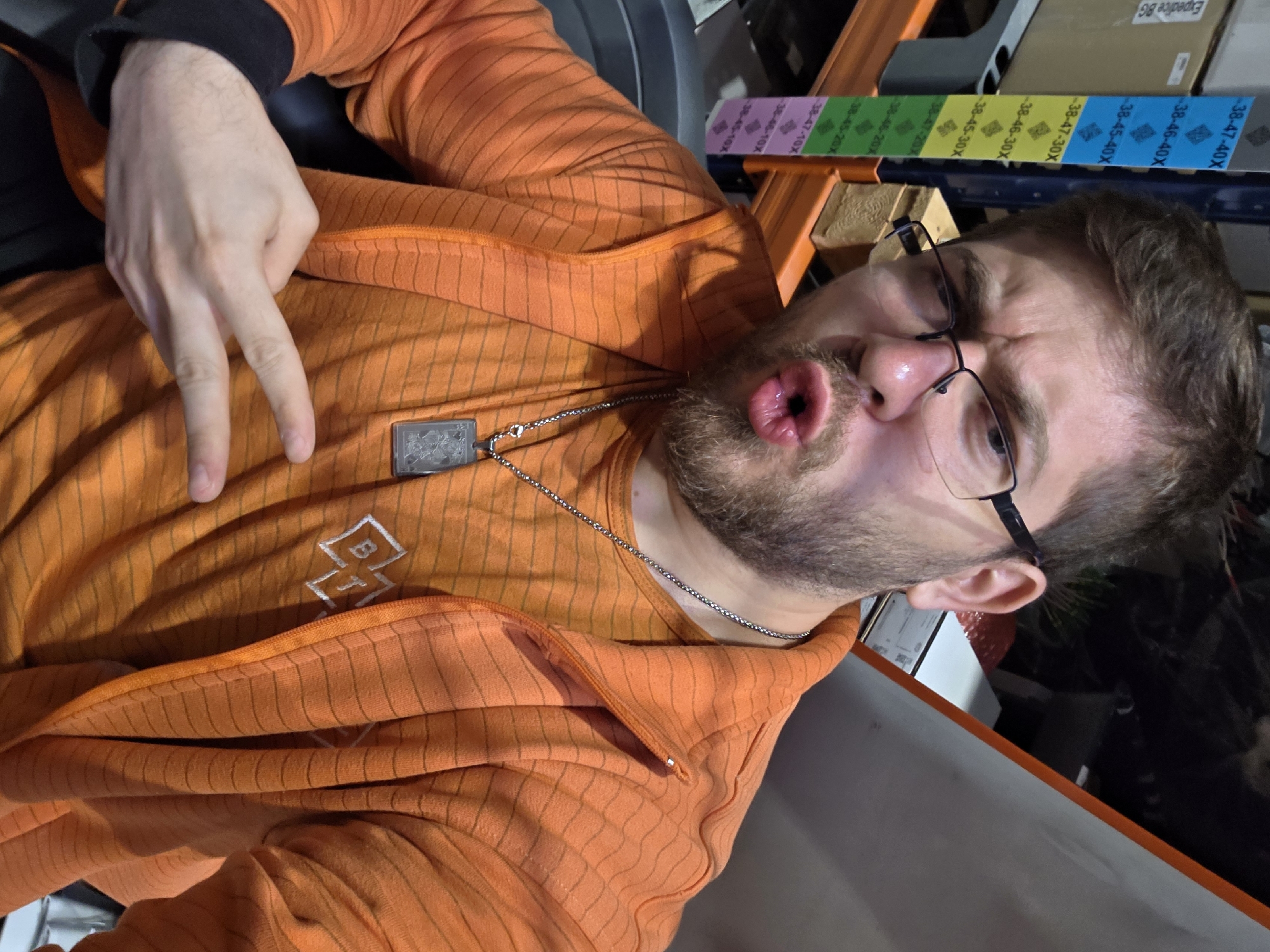 A man is pictured indoors, close to a shelving unit with size labels attached. The man wears an orange zip-up hoodie and a necklace with a square pendant. He is making a duck-face expression and holding up two fingers in a peace sign. He has short brown hair, a beard, and wears glasses. The background shows shelving with labels and storage boxes. The location cannot be confirmed in Plovdiv, Bulgaria, based on visual evidence.