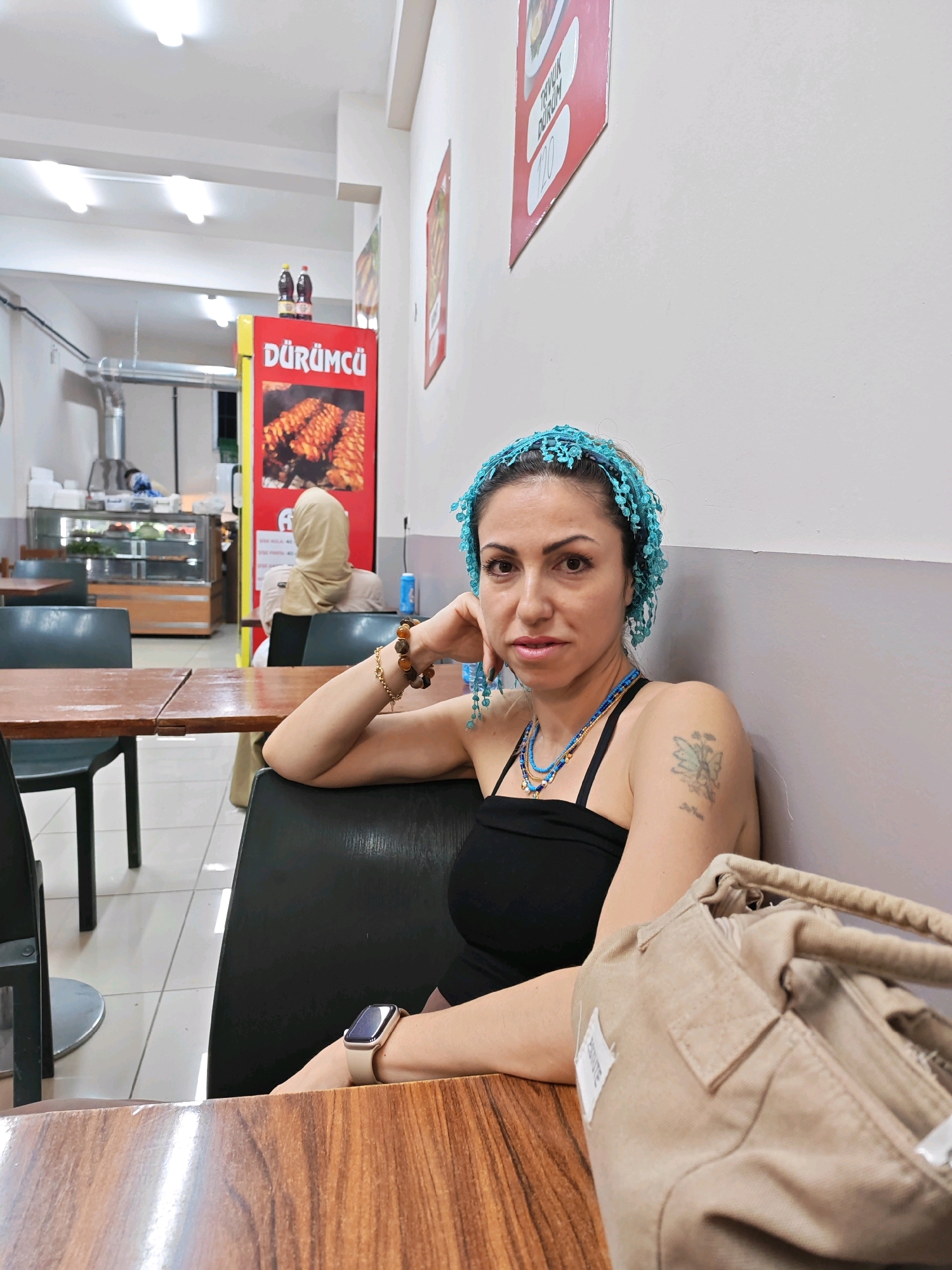 In Zeytinburnu, Turkey, a woman with a blue headscarf and a black tank top sits at a wooden table in a restaurant. She has her left arm resting on her lap and her right hand on her cheek. A beige handbag rests on the table in front of her. In the background, a red sign displays pictures of grilled meat and the word "DÜRÜMCÜ". Another person, wearing a beige headscarf, is seated at a table further back. The restaurant appears to be clean and well-lit, with a display case containing food items visible.