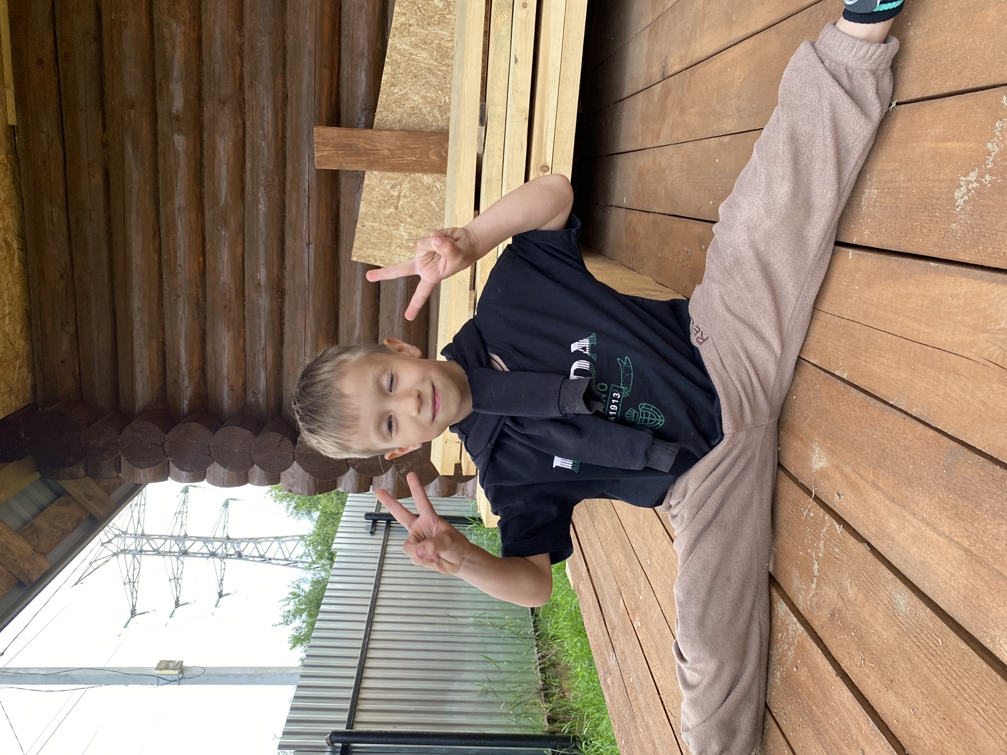A young boy with blond hair, wearing a black t-shirt and beige sweatpants, is pictured on a wooden deck. He is smiling and making a peace sign with his left hand. His legs are spread in a split, with his right leg extended forward and his left leg bent. The background shows a wooden structure, possibly a porch or gazebo, with visible logs and planks. Beyond the wooden structure, a metal fence and power lines are visible against a bright sky. The scene appears to be outdoors, with natural light suggesting daytime. The boy's pose and expression convey a sense of playfulness and cheerfulness. Visible text on his t-shirt includes the words "RE" and "1913".