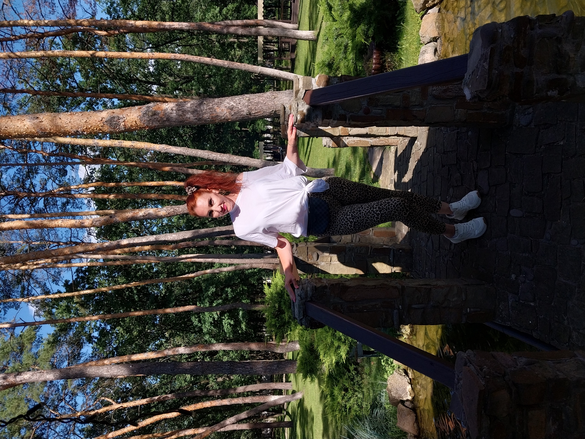 A woman with red hair in a ponytail stands on a stone bridge in a park. She is wearing a white off-the-shoulder top tied at the waist and leopard-print leggings. She has on white sneakers and a small black fanny pack. The bridge has stone pillars and wooden railings. To the left of the bridge is a small pond with calm, clear water and rocks along its edge. To the right of the bridge is a grassy area with a few bushes. Behind the bridge, a dense forest of tall pine trees with sparse green foliage creates a backdrop. The sun is shining, casting shadows on the bridge and the surrounding area. The overall impression is a sunny day in a natural, wooded setting.
