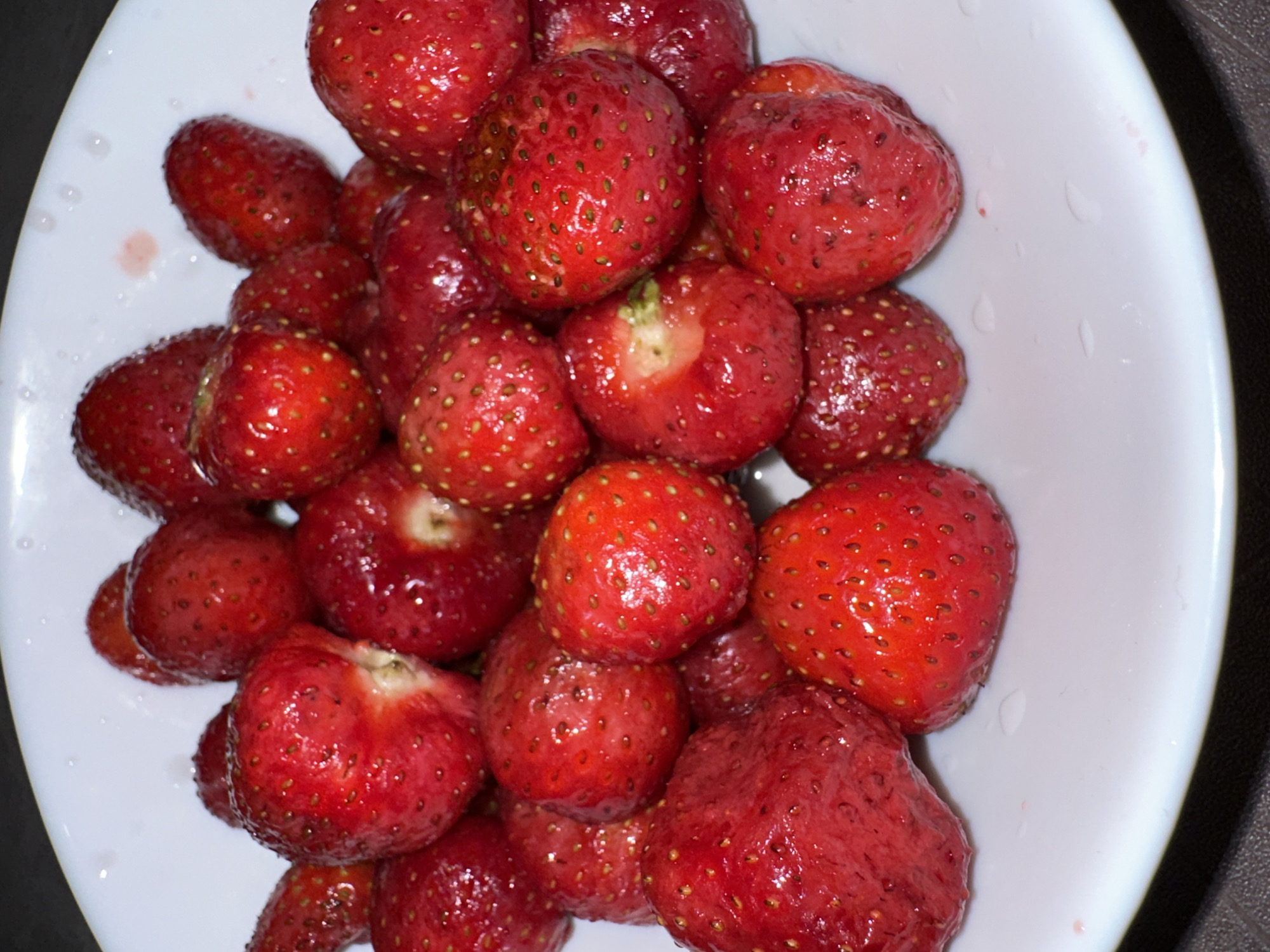 A close-up, overhead shot displays a white bowl filled with ripe strawberries. The strawberries are a vibrant red, with some showing small seeds on their surface. A few of the strawberries have been bitten into, revealing their pale green interior. The bowl is placed on a dark, textured surface. No specific location in Ternopil, Ukraine can be determined from this image.