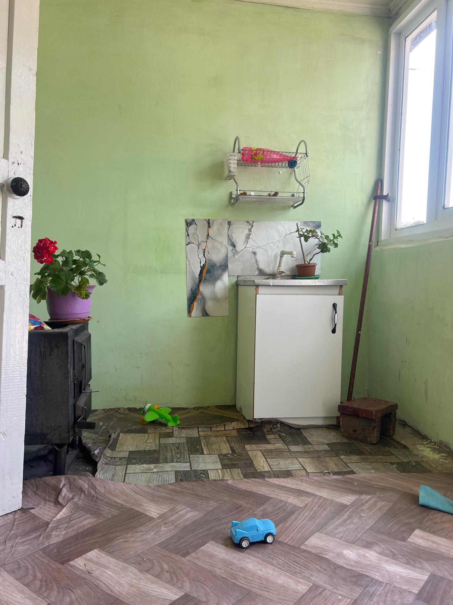 An interior scene, likely a kitchen or utility area, is captured in a dimly lit room. The walls are painted a light green, and the floor is covered in a patterned vinyl material that appears to be peeling in some areas.
In the left foreground, a dark-colored wood-burning stove is partially visible, with a potted red geranium placed on top of it. Adjacent to the stove, a white door frame is present.
The central area of the room features a white cabinet with a black handle, topped with a countertop that has a faux marble backsplash. A small potted plant sits on the countertop. To the right of the cabinet, a metal rod leans against the wall, and a small wooden stool is positioned nearby.
The background includes a window with white framing, through which daylight is entering. A metal dish rack with some items on it is mounted on the wall above the cabinet. On the floor, scattered toys are visible, including a green toy airplane and a blue toy car. The overall impression is one of a functional but somewhat unkempt space. The location is identified as Dzhida, Kyrgyzstan.