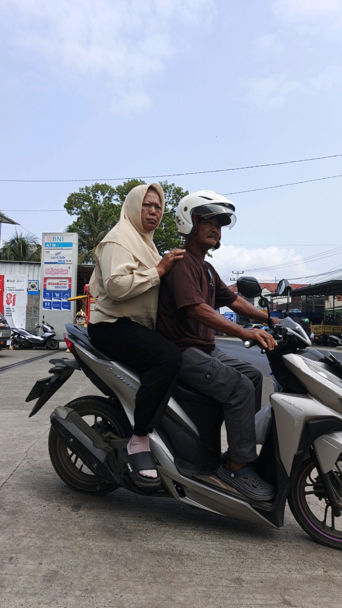 In Pangandaran, Indonesia, a man and a woman ride a silver Honda motorcycle. The man, wearing a brown shirt and gray pants, drives the motorcycle, his right hand on the handlebar. He has a mustache and wears a white helmet. The woman, in a beige hijab and a light-colored shirt, sits behind him, her left hand resting on his shoulder. She wears black pants and black slides. The motorcycle is parked on a concrete surface, with a gas station visible in the background. The sky is partly cloudy.