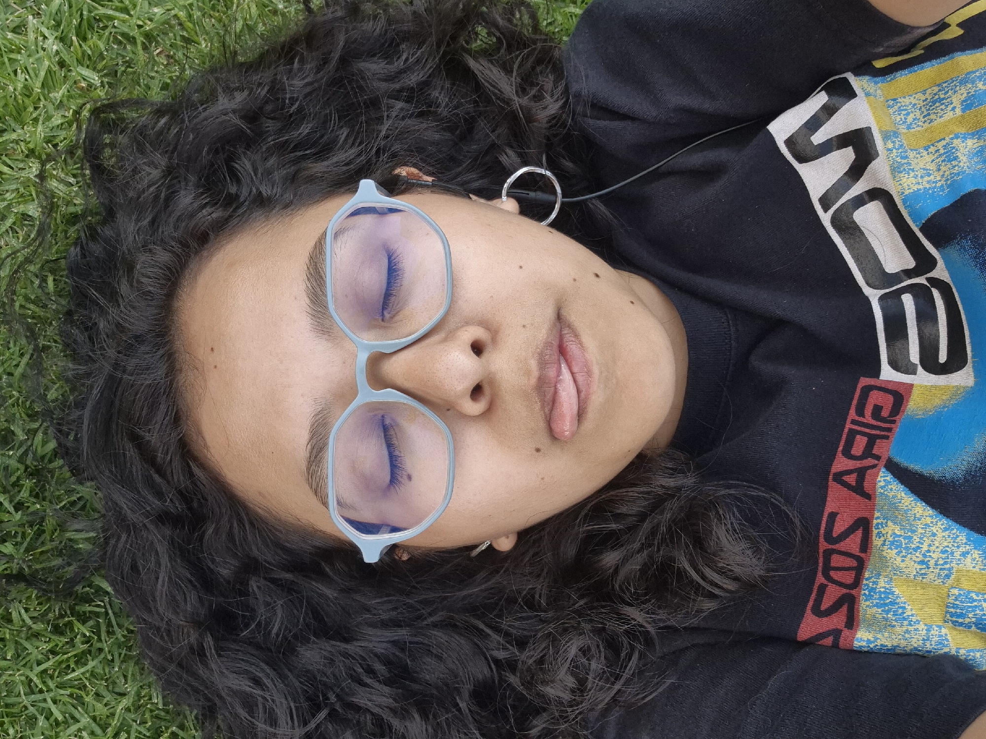 A woman lies on her back in the grass, wearing light blue glasses. Her eyes are closed, and her mouth is slightly open, revealing the tip of her tongue. She has dark, curly hair that spreads around her head on the grass. She wears a black t-shirt with a graphic design featuring the word "ASOS" in red on the right side. A silver earring is visible in her left ear, along with an earbud. The location appears to be a grassy area, possibly a park, but it is not possible to determine the exact location in Puebla, Mexico, from the image alone.